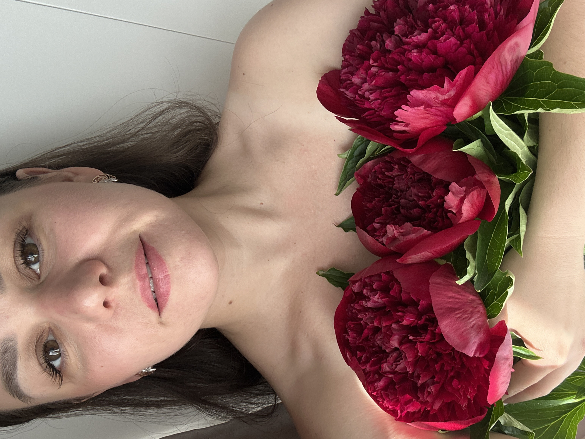 The image, set in Chaikovsky, Russia, features a woman lying down, angled towards the viewer with her head on the left side of the frame. She has long, dark brown hair, brown eyes, and light skin, with visible bare shoulders and upper chest. Her lips have a subtle pink tint, and her eyes are accentuated with mascara. She wears small, intricate metallic earrings in both ears. Alongside her, three large, vibrant dark red peonies with prominent green leaves are clustered against her body, partially obscuring her right arm and torso. The woman gazes directly at the camera with a soft, faint smile, conveying a calm and serene emotion. The background is a plain, light, neutral color, possibly a wall or bedsheet, indicating an indoor setting. The image is oriented such that the woman appears to be lying on her back, viewed from above, with the entire composition rotated approximately 90 degrees clockwise. There is no visible text in the image.