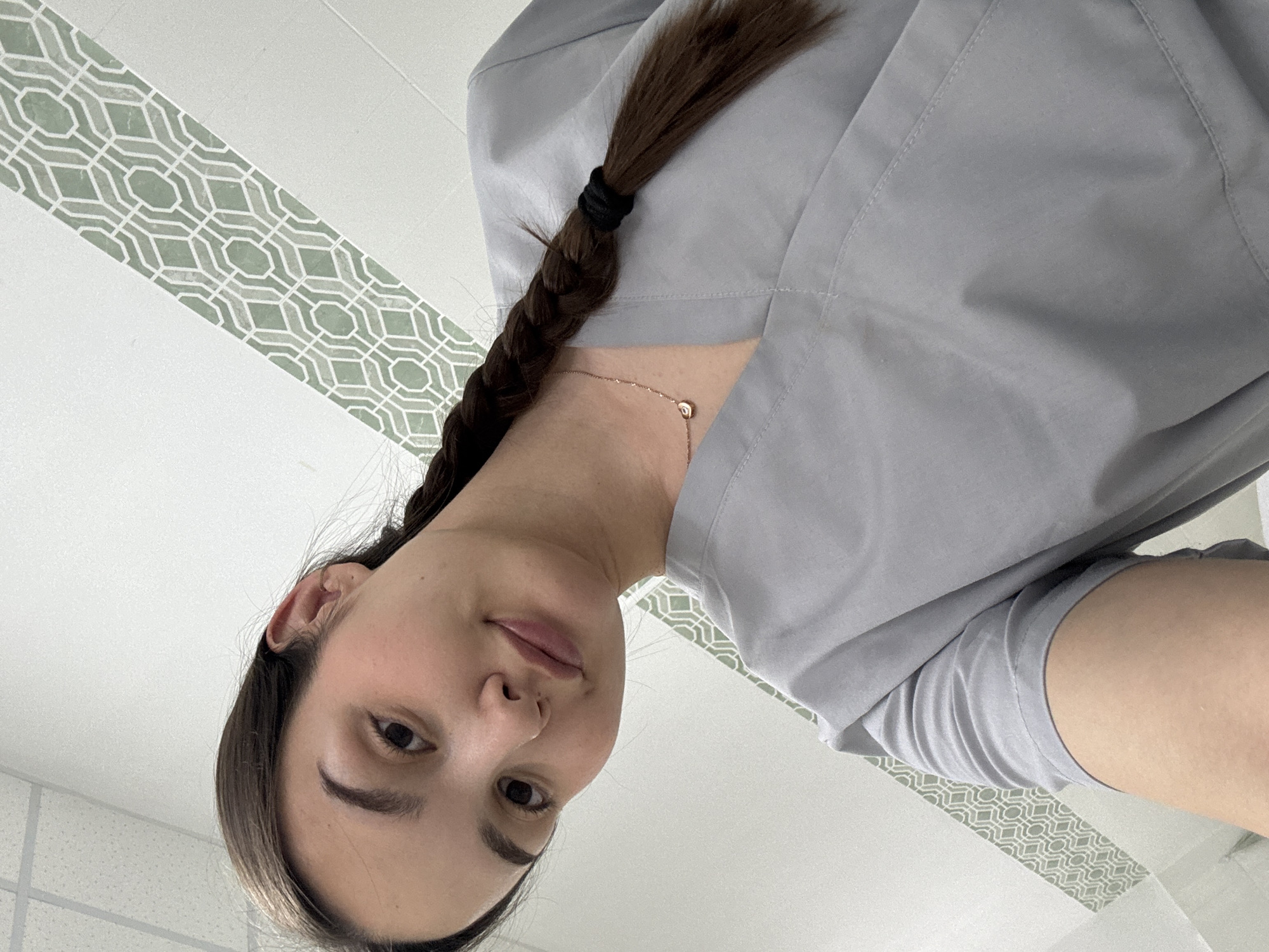 A young woman is featured in a close-up, diagonally oriented selfie, looking directly at the camera with a neutral to subtly smiling expression. Her head is tilted, and she has fair skin, dark eyebrows, and long dark brown hair styled in a neat braid, secured with a black elastic, falling over her right shoulder. She is dressed in a light grey uniform top, possibly scrubs, which has visible seam details. A delicate gold-toned necklace with a small, round pendant is visible around her neck.
The background suggests an indoor setting, primarily white. A prominent decorative feature is a wide diagonal stripe across the background, displaying a repetitive geometric pattern in light green and white/light grey, reminiscent of a trellis or mosaic design. In another section of the background, a grid pattern consistent with acoustic ceiling tiles can be seen. The lighting is bright and even, typical of an interior space. Her attire implies a professional environment, potentially within a medical or service industry in Chaikovsky, Russia. No text is visible in the image.