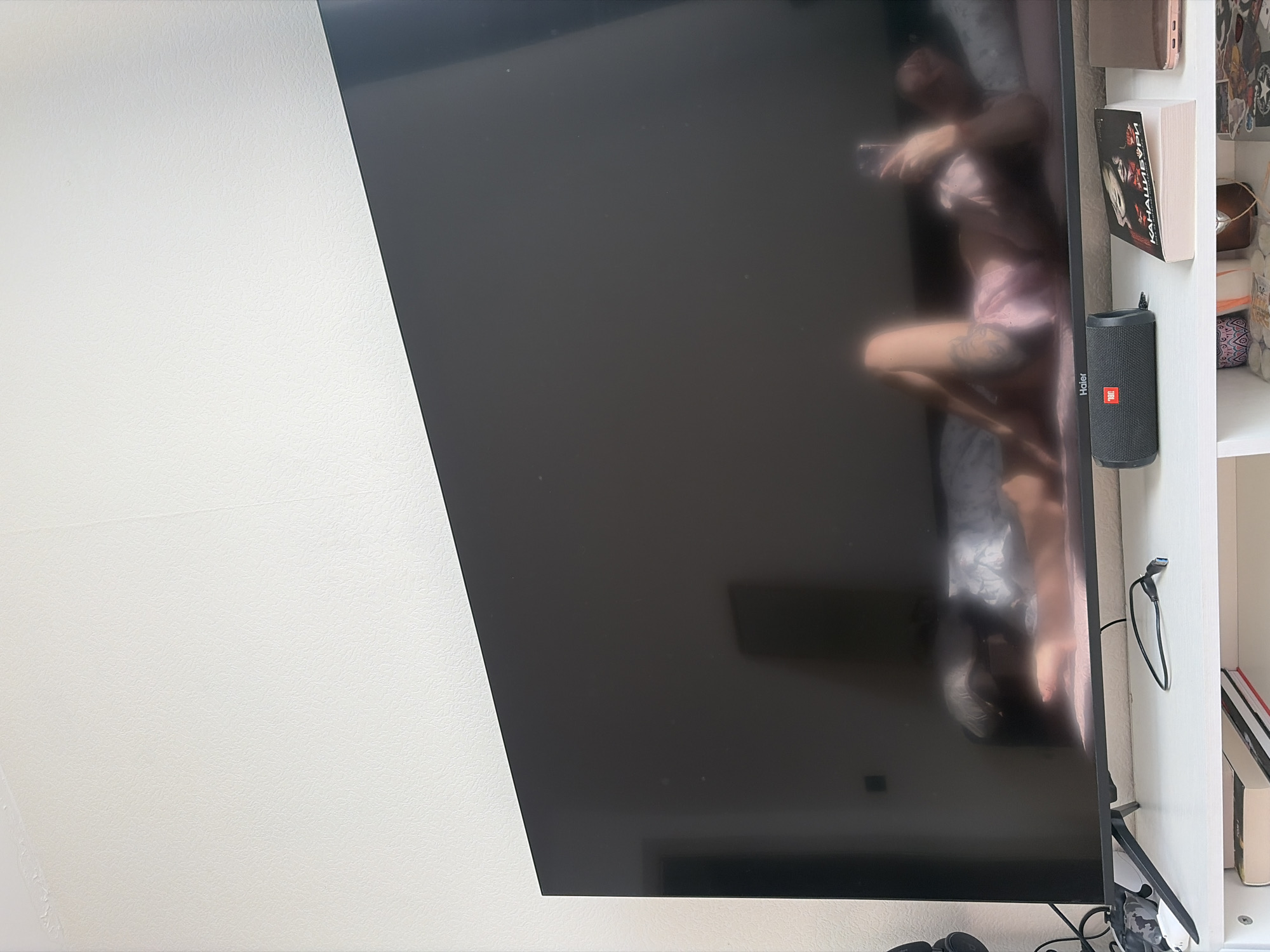 A television screen reflects a woman in pink pajamas holding a mobile phone, standing or lying on a bed. The television is positioned next to a white bookcase containing books, a JBL speaker, and other items. The scene is indoors, with a white wall visible to the left of the television. There is no confirmed evidence the image was taken in St. Petersburg.