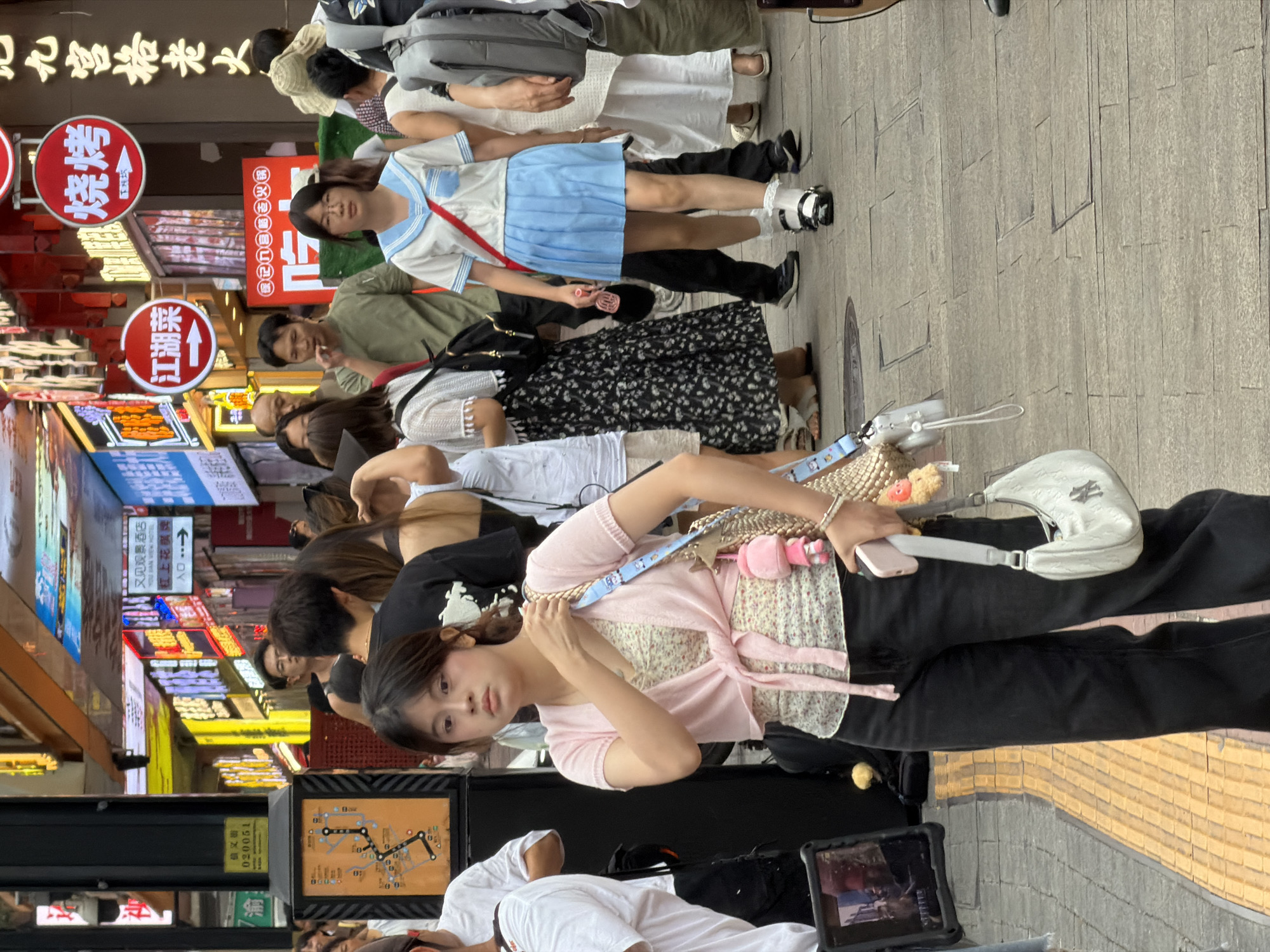 In this outdoor scene, several individuals are present, with a young woman in the foreground drawing particular attention. She is wearing a light pink cardigan over a floral top and black pants, and holding a pink phone. Behind her, other people are visible, including one in a school uniform with a blue skirt and white top. The setting appears to be a bustling street, possibly a shopping area, judging by the numerous neon signs in the background, many of which display Chinese characters. Some of the visible text on the signs includes "红街" (Red Street) and "又见龙泉" (See Longquan Again). The ground is paved with stone tiles. The lighting suggests it is daytime, and the overall atmosphere seems relaxed and ordinary, with people going about their day.