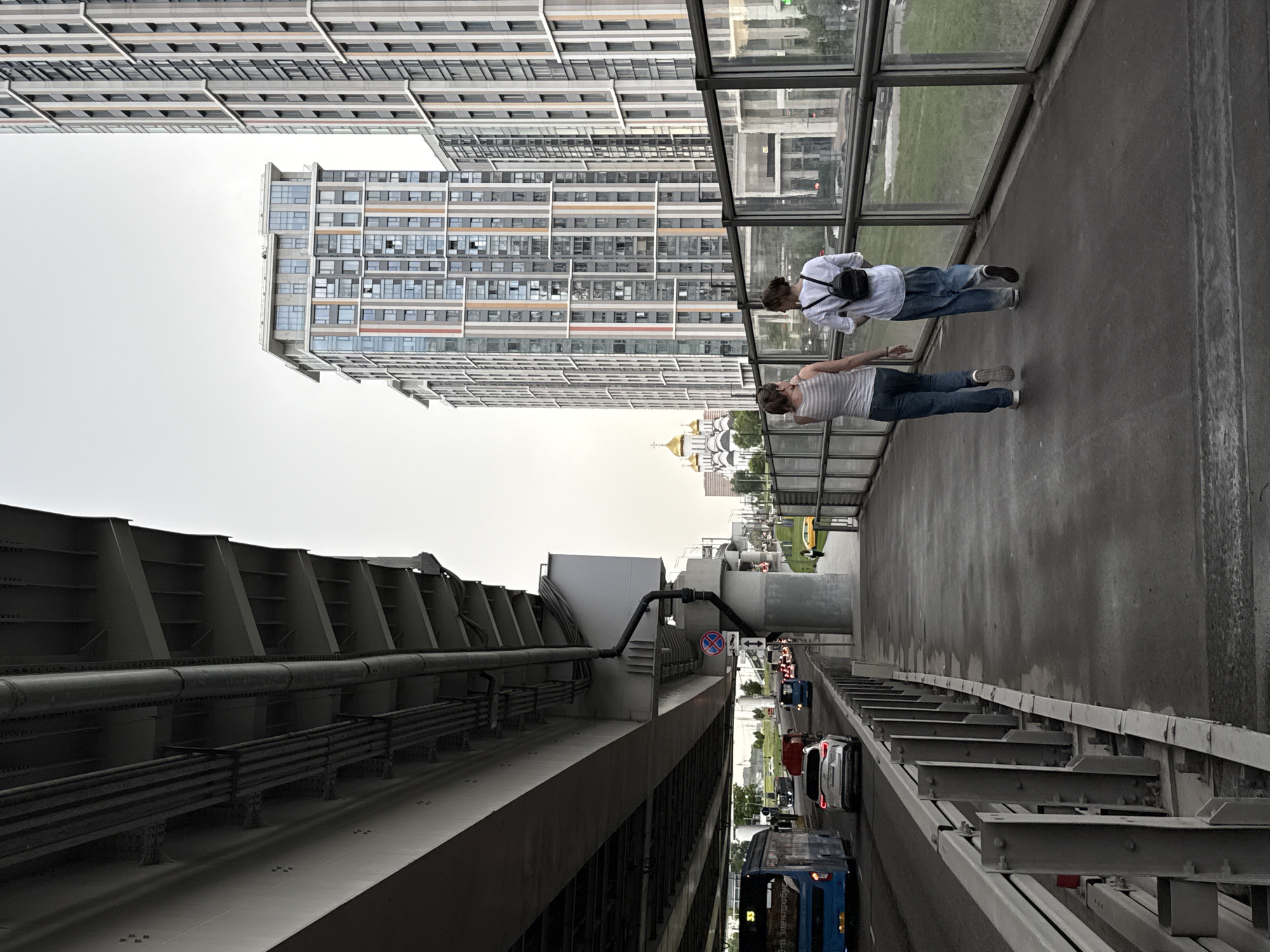 The image shows a wide pedestrian walkway overlooking a busy street in Moscow, Russia. The walkway is separated from the street by a glass barrier and a metal railing. Two young women are walking away from the camera on the walkway. The woman on the left is wearing a striped tank top and jeans, while the woman on the right is wearing a white t-shirt, jeans, and a black backpack.
In the background, a modern multi-story building with a distinctive facade featuring vertical bands of color stands tall. To the left of this building, a church with golden domes is visible. The street below is lined with cars and buses, suggesting a bustling urban environment. A large elevated highway structure is prominent in the foreground, casting a shadow over the street and walkway. The sky is overcast.