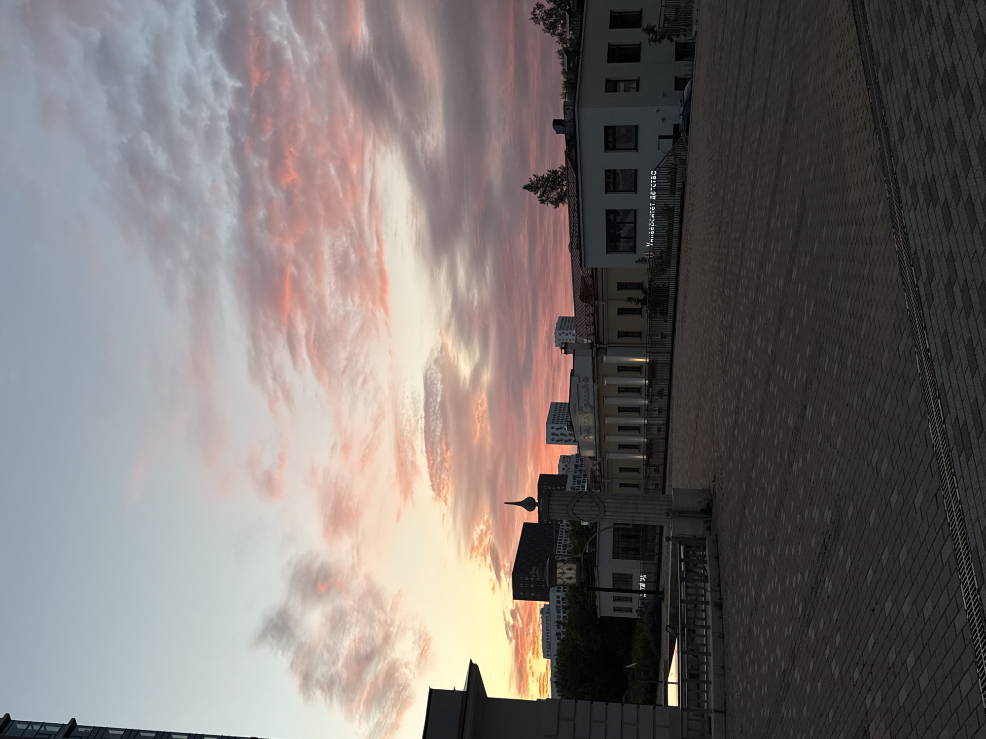 The image depicts a public square in Moscow, Russia, at dusk. The sky is filled with dramatic pink and orange clouds, indicating the setting sun. In the background, several modern buildings of varying heights and architectural styles are visible, including a dark, rectangular high-rise. The foreground features a paved area with a patterned brick surface and a metal grate. A low metal fence runs along the edge of the paved area. A stone pillar with a decorative finial stands prominently in the mid-ground. A building with the text "Университет детства" (University of Childhood) in Russian is visible on the right side. No people are present in the scene.