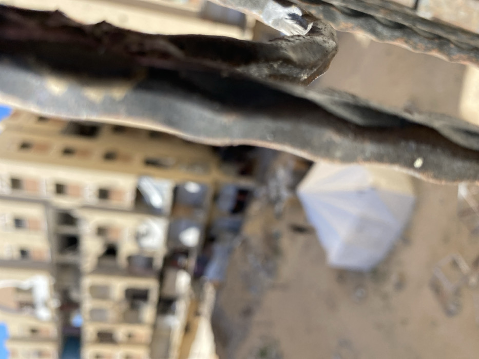 An elevated, downward-angled view captures a scene of extensive urban destruction in Khan Yunis, Palestinian Territory, through a blurred foreground element. The immediate foreground on the right displays an out-of-focus, dark, possibly metallic or wrought iron structure, suggesting part of a balcony or railing. The midground and background depict multiple multi-story buildings, predominantly light brown or beige, exhibiting severe structural damage with large sections of walls missing and interiors exposed. Below these damaged structures, an open, sandy or dusty ground area contains scattered rubble. A prominent white, peaked tent structure is situated on this ground, slightly left of the center. Indistinct, small figures are discernible on the ground near the tent and damaged buildings. A clear blue sky is visible above the horizon. The entire distant scene appears under daylight conditions.