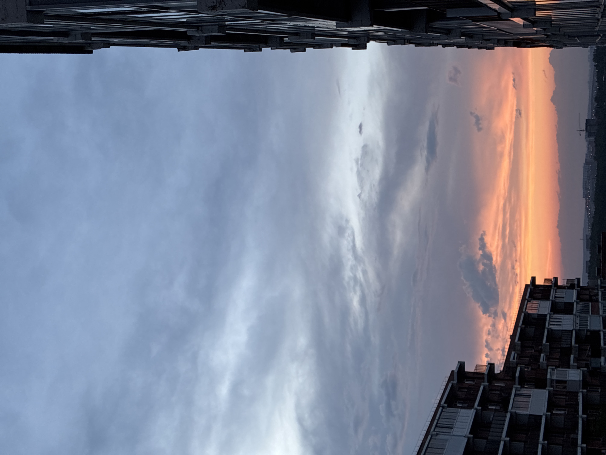 An exterior, eye-level, wide shot captures a twilight cityscape in Markova, Russia. The sky is filled with soft, scattered clouds in hues of blue, gray, and orange. Two large apartment buildings dominate the foreground, one on the left and the other on the right, casting long shadows. The building on the left is darker, with many windows and balconies. The building on the right is lighter, with a concrete exterior and rows of windows. In the distance, a cluster of smaller buildings and a construction crane are visible. The overall atmosphere is serene and peaceful.