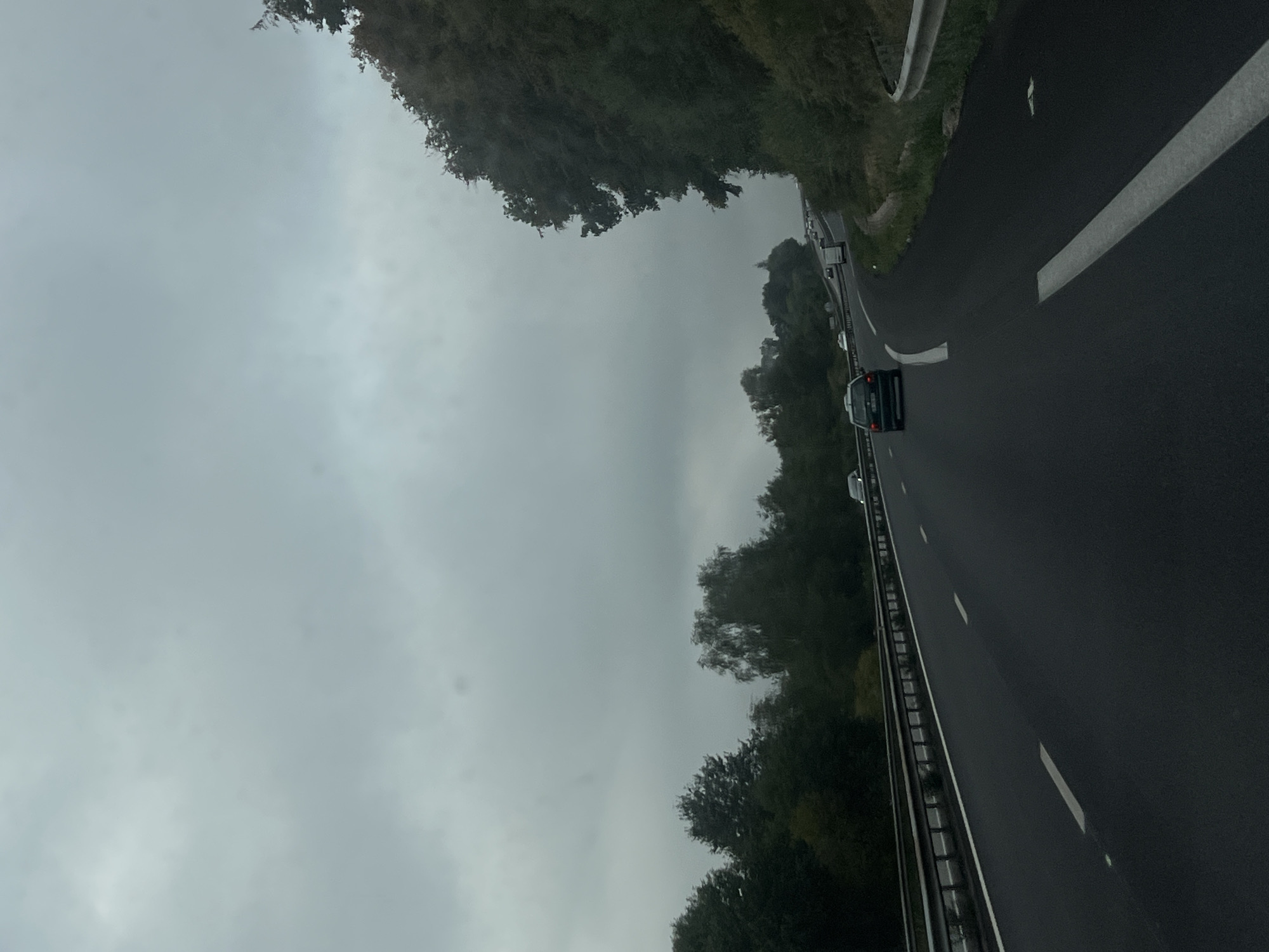 The image shows a view from a vehicle traveling on a highway in Razès, France. The sky is overcast with dark, heavy clouds. The highway has multiple lanes, indicated by white dashed lines. A guardrail separates the highway from the shoulder and embankment. On the left side of the highway, there is a dense line of trees and foliage. On the right side, the embankment is also covered with trees and vegetation, with a guardrail visible. Several vehicles are on the highway, including a dark-colored car in the foreground in the right lane, and other vehicles further ahead. The overall scene suggests a gloomy, possibly rainy, day.