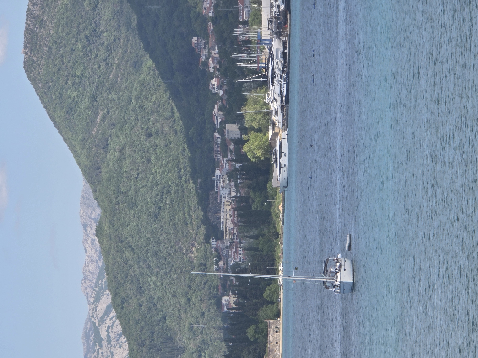 The image shows a view of the coast in Đuraševići, Montenegro. A large, forested mountain rises on the left side of the frame. A small town is situated along the coast at the base of the mountain, with buildings interspersed among trees. A marina with numerous boats is visible along the waterfront. A single sailboat is anchored in the blue water of the bay. The sky is clear.