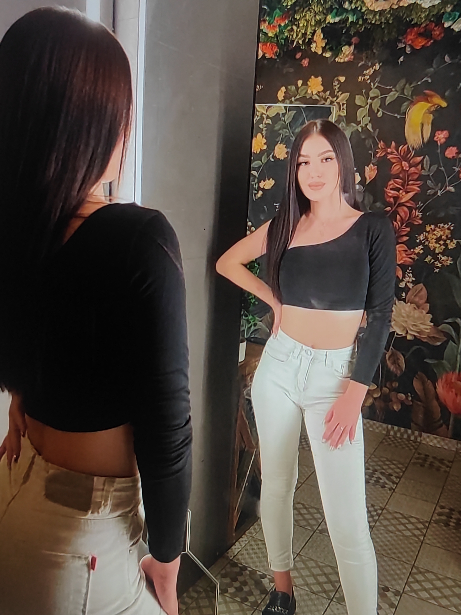 The image shows a young woman posing in front of a floral-patterned wallpaper. She is wearing a black, one-shoulder crop top and white skinny jeans. Her dark hair is long and straight. The woman is standing in what appears to be an indoor setting, possibly a room with a mirror and a tiled floor. The wallpaper features a colorful botanical design with birds and flowers. She is looking towards the camera, with one hand on her hip. The lighting suggests it could be daytime or evening. The overall mood is stylish and confident. There is no visible text in the image.