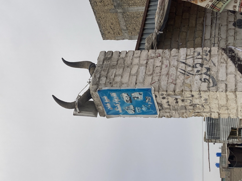 The scene depicts an outdoor environment in Quetta City Tehsil, Pakistan, under an overcast, light grey sky. In the foreground, slightly right of center, stands a weathered brick pillar, appearing to be constructed from light-colored or whitewashed bricks, displaying areas of dirt and dark graffiti. Mounted atop the pillar are two large, dark, curved animal horns, secured with visible wires or straps. A rectangular light fixture is affixed to the front of the left horn structure. Below the horns, a blue banner or poster is attached to the pillar's front face. This banner features Urdu text at the top, translatable as "Nero Khan Car Parking," followed by six white graphic icons depicting various vehicles: a large truck, a car, a three-wheeled auto-rickshaw, a cement mixer, a motorcycle, and another three-wheeled utility vehicle. To the immediate right of the pillar, a structure with a corrugated metal roof is partially visible, with an adjacent building constructed of light brown bricks appearing in the background. Further left in the mid-ground, additional structures with corrugated metal roofs and blue water storage tanks on their rooftops are visible. A small section of a checkered fabric is partially visible in the bottom right corner of the frame. No individuals are present in the image.