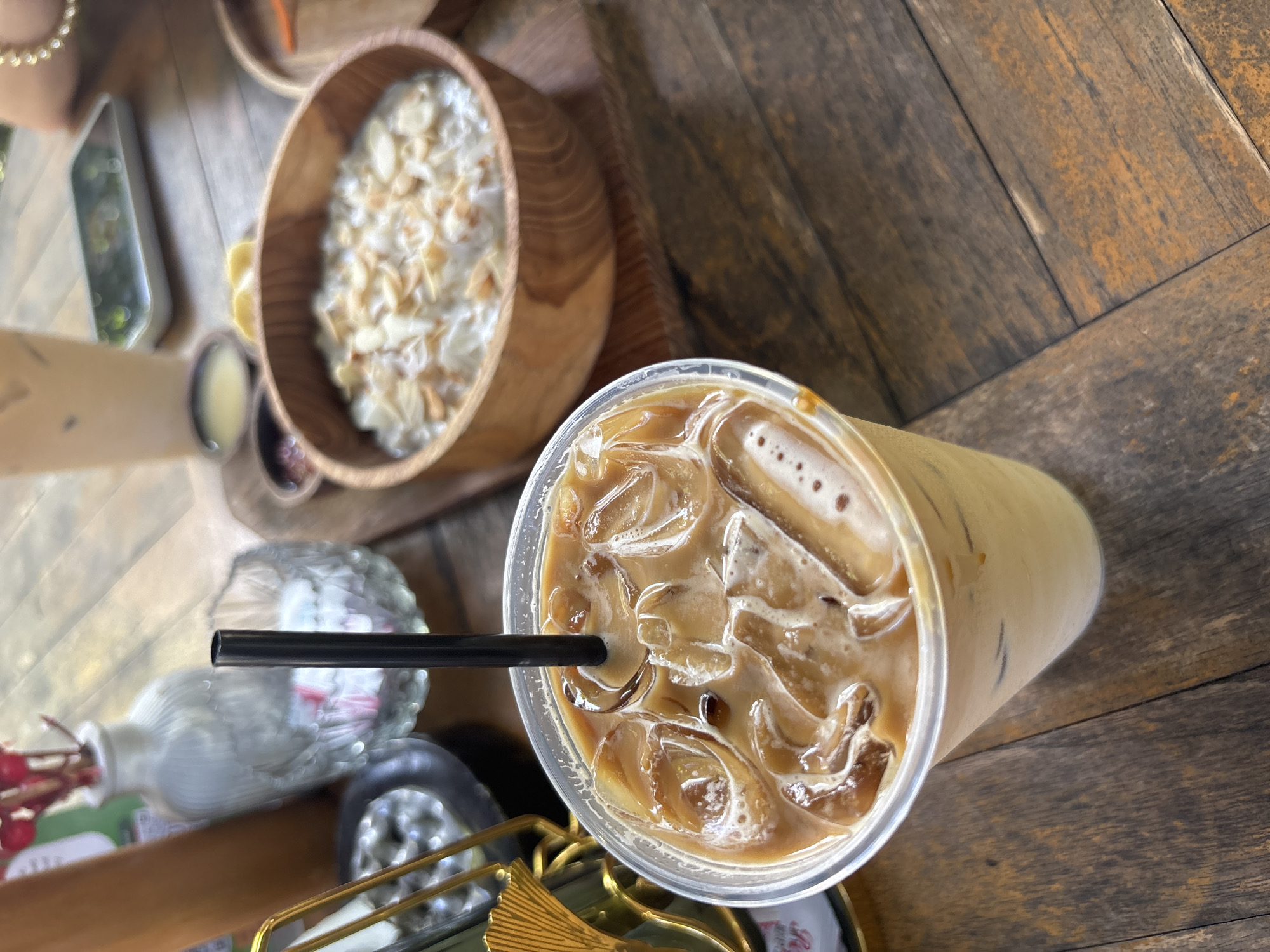 On a wooden table in an establishment in Kuta Selatan, Indonesia, sits a breakfast spread. A wooden bowl contains a white, creamy substance with light-colored sliced nuts on top. Next to it, a clear plastic cup holds an iced coffee with a black straw inserted. Further to the left, a small white vase with red stems and green leaves is visible, along with miscellaneous tableware. The table's wooden surface has a distinct grain pattern, indicating a rustic, natural environment.