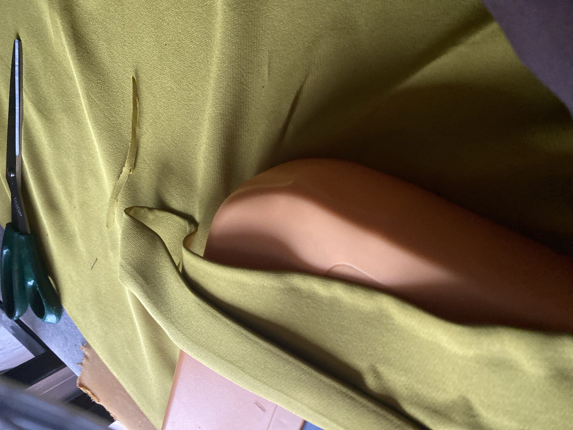 The image shows a close-up view of a light green fabric draped over an orange object, possibly a mannequin or a body form. A pair of green-handled scissors lies on the fabric. There are also some darker markings or shadows on the fabric, as well as a frayed edge of a lighter colored material in the background. The lighting suggests an indoor setting. The image is cropped in a way that makes it difficult to determine the overall context or environment. There is no indication of the city of Jalingo, Nigeria, or any specific location within it.
