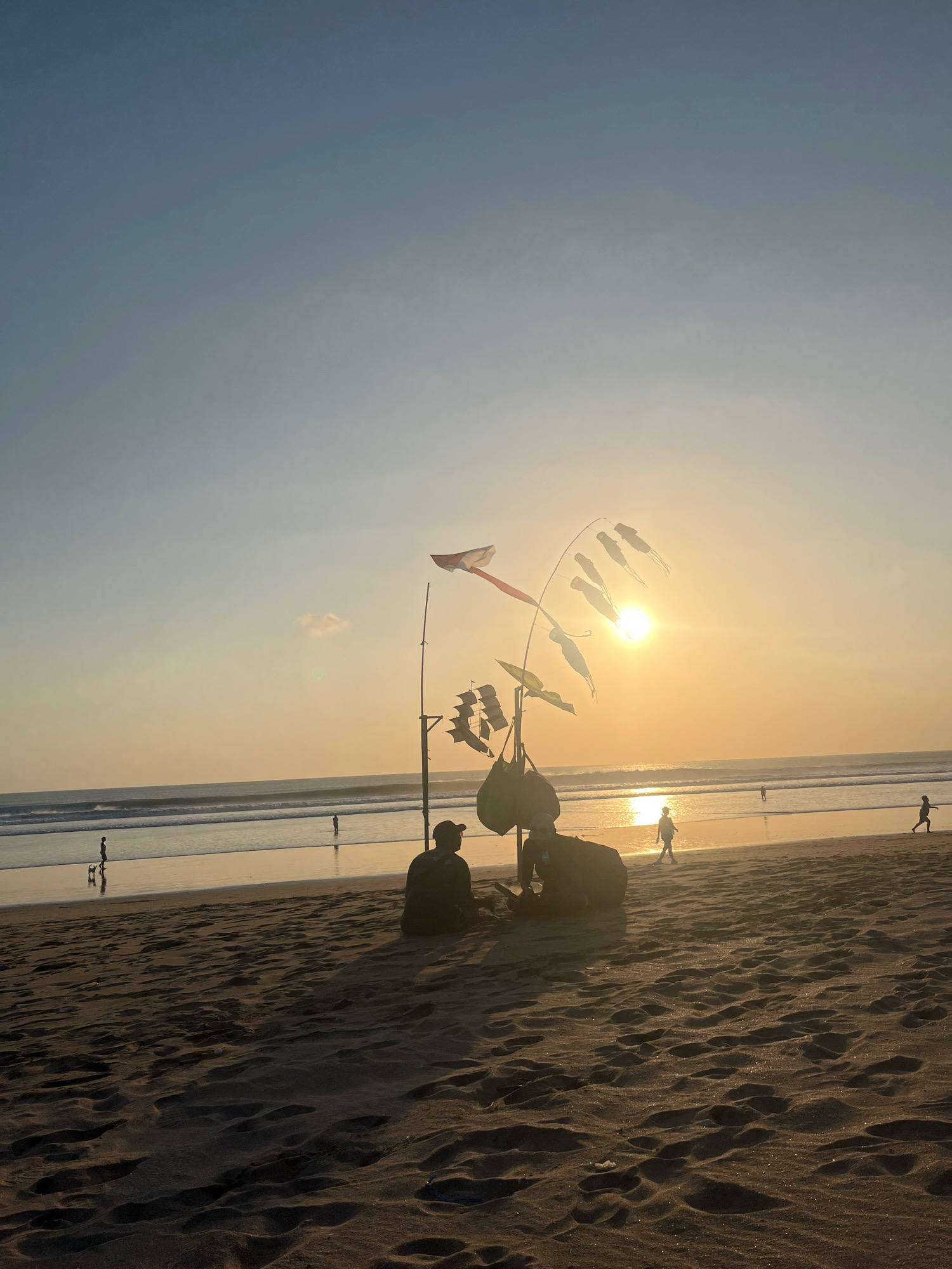 The image depicts a beach scene in Kuta, Indonesia, during sunset. The sun is low on the horizon, casting a warm glow across the sky and water. Several people are visible on the beach, some walking along the shoreline, while others are sitting on the sand. In the foreground, two figures are seated, with a tall bamboo pole erected nearby, adorned with decorative flags or kites shaped like fish and a ship. The waves are gently rolling onto the sandy shore, reflecting the golden light of the setting sun. The sky is a gradient of pale blue and orange, with a few wispy clouds. The sand is textured with footprints and patterns left by the receding tide.
