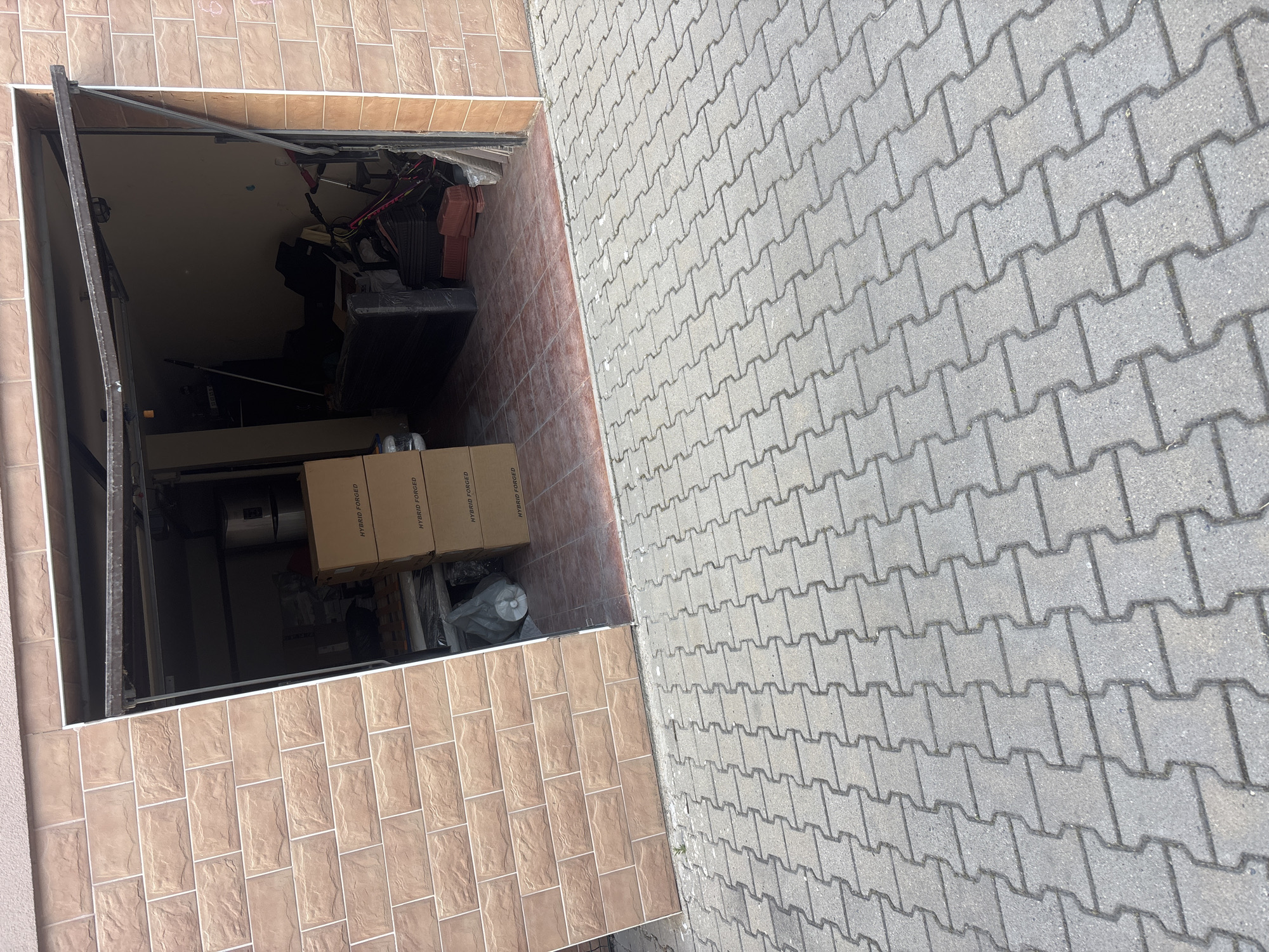 The image shows a garage opening onto a gray, brick-paved area. The garage interior is cluttered with various items, including boxes with the label "HYBRID FORGED". The garage walls and trim are tiled in a light brown, textured brick. There are no people visible. The location is unknown, but specified as Plovdiv, Bulgaria.