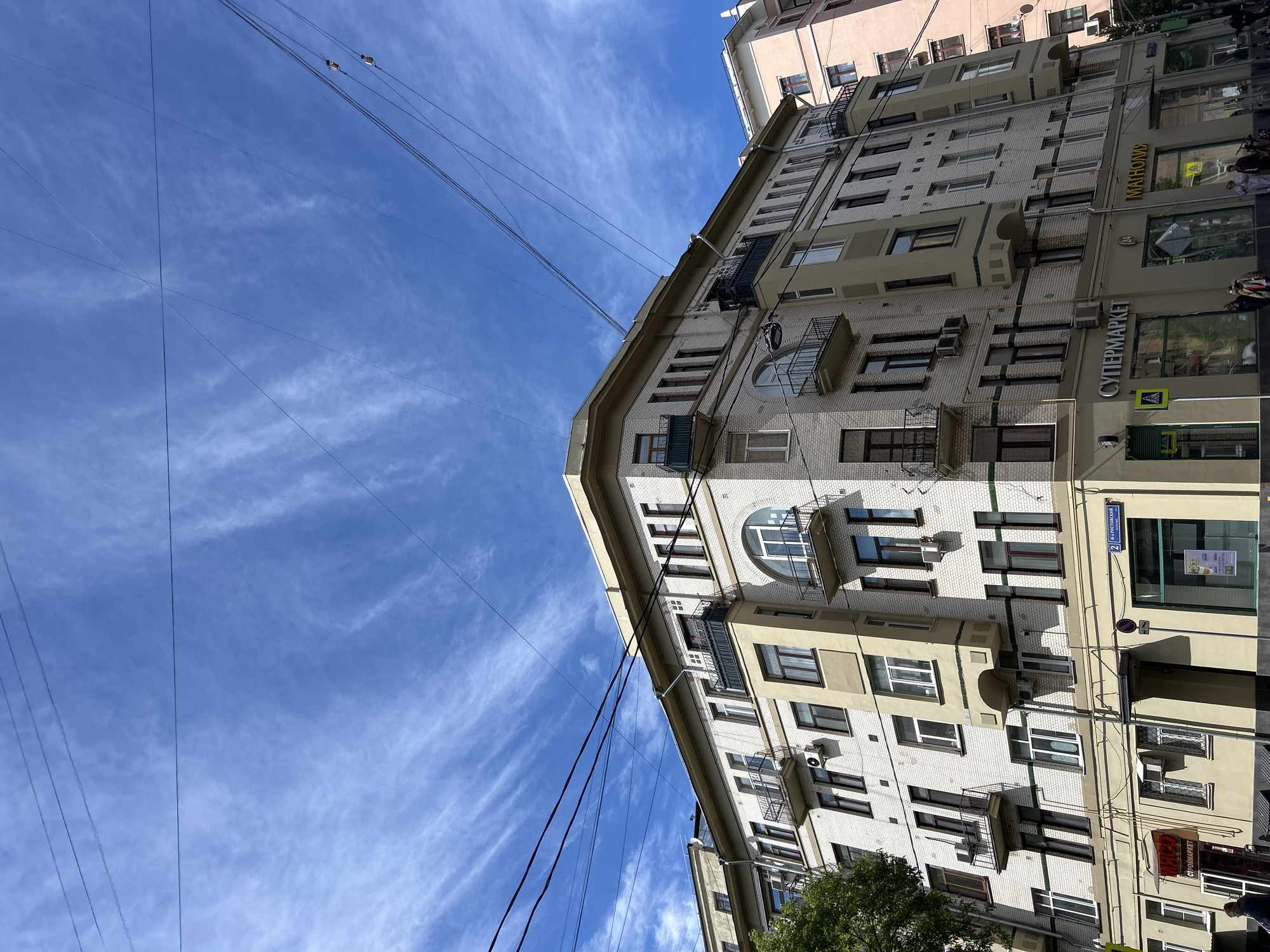 The image captures an urban scene in an unspecified city. Dominating the composition is a tall, light-colored building with multiple stories and a distinctive architectural style. The building's facade features a mix of white and tan elements, with numerous windows and small balconies. Several air conditioning units are affixed to the exterior walls. Signage in a non-English language indicates that the building houses commercial establishments, including a "CУПЕРМАРКЕТ" (Supermarket). A pink building is visible directly above the light-colored building. Overhead, power lines intersect the blue sky, which is partially covered by wispy cirrus clouds. The camera angle is tilted upwards, emphasizing the height of the buildings and the expansive sky above.