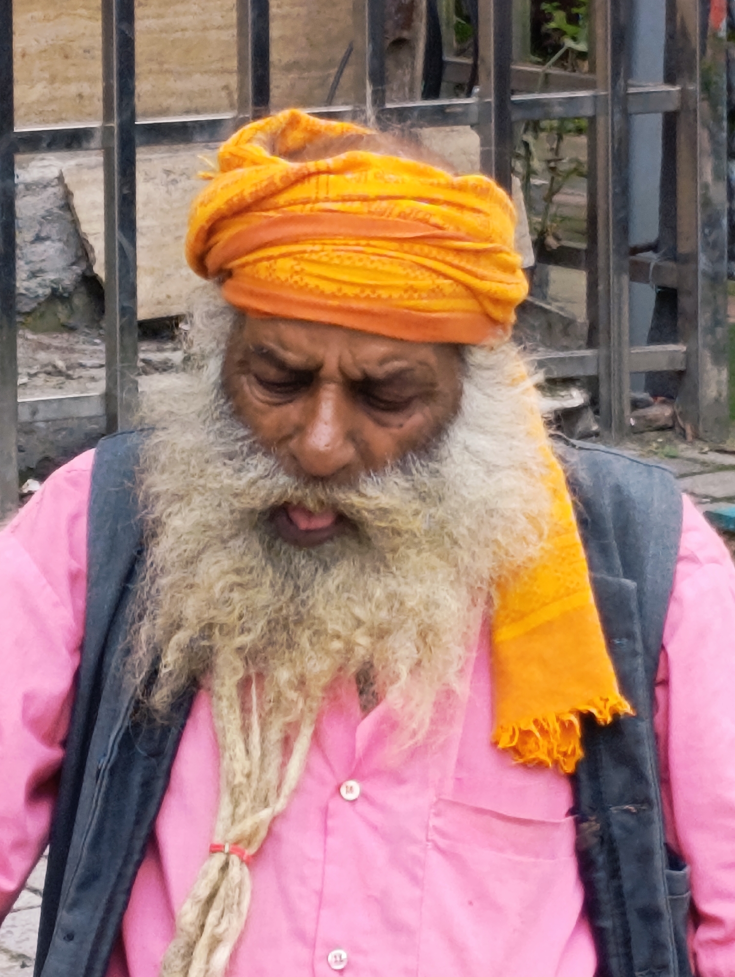 A close-up, eye-level, portrait-oriented, medium shot of an elderly man, likely a Sadhu, in Manali, India, features him with his head turned slightly down and to the right. He wears a bright orange turban wrapped around his head and a pink collared shirt with buttons visible. A dark grey vest is worn over the shirt. His long, thick, grey-blonde beard is prominent and reaches his chest, with several strands of his hair braided and tied with a red band. He has dark skin, a dark eyebrow, and a mustache. His mouth is slightly open, revealing his tongue. An orange scarf with a tasseled edge is draped around his neck, with one end hanging over his right shoulder. The background consists of a dark metal fence and a weathered, light-colored wall or structure. The lighting is even, suggesting an overcast day or shade.