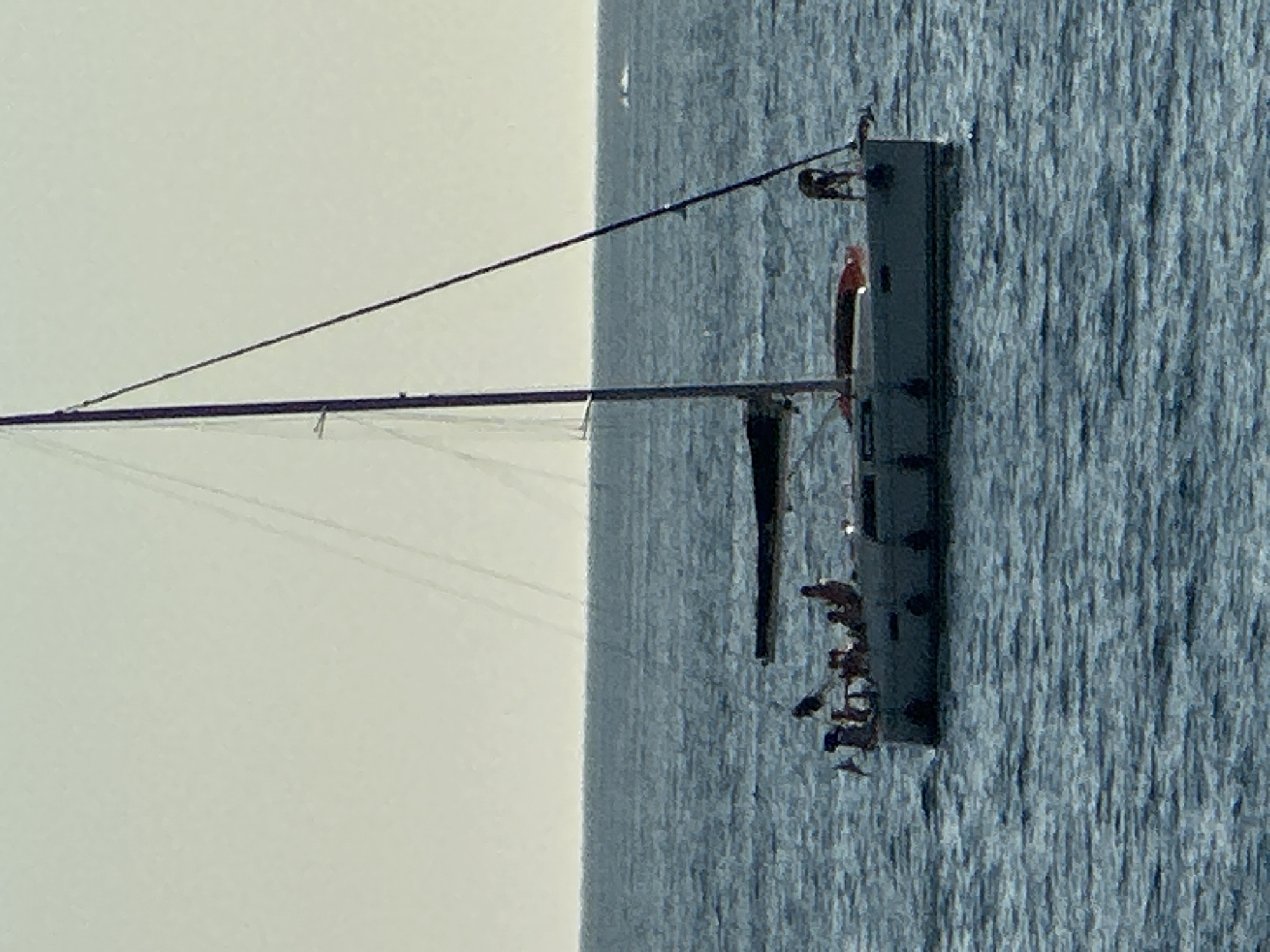 A sailboat with several individuals on its deck is on the water. The boat has a dark sail, a mast, and rigging. The water is choppy and blue, and the sky is a light, hazy color. In the distance, a small boat with a white wake is visible.