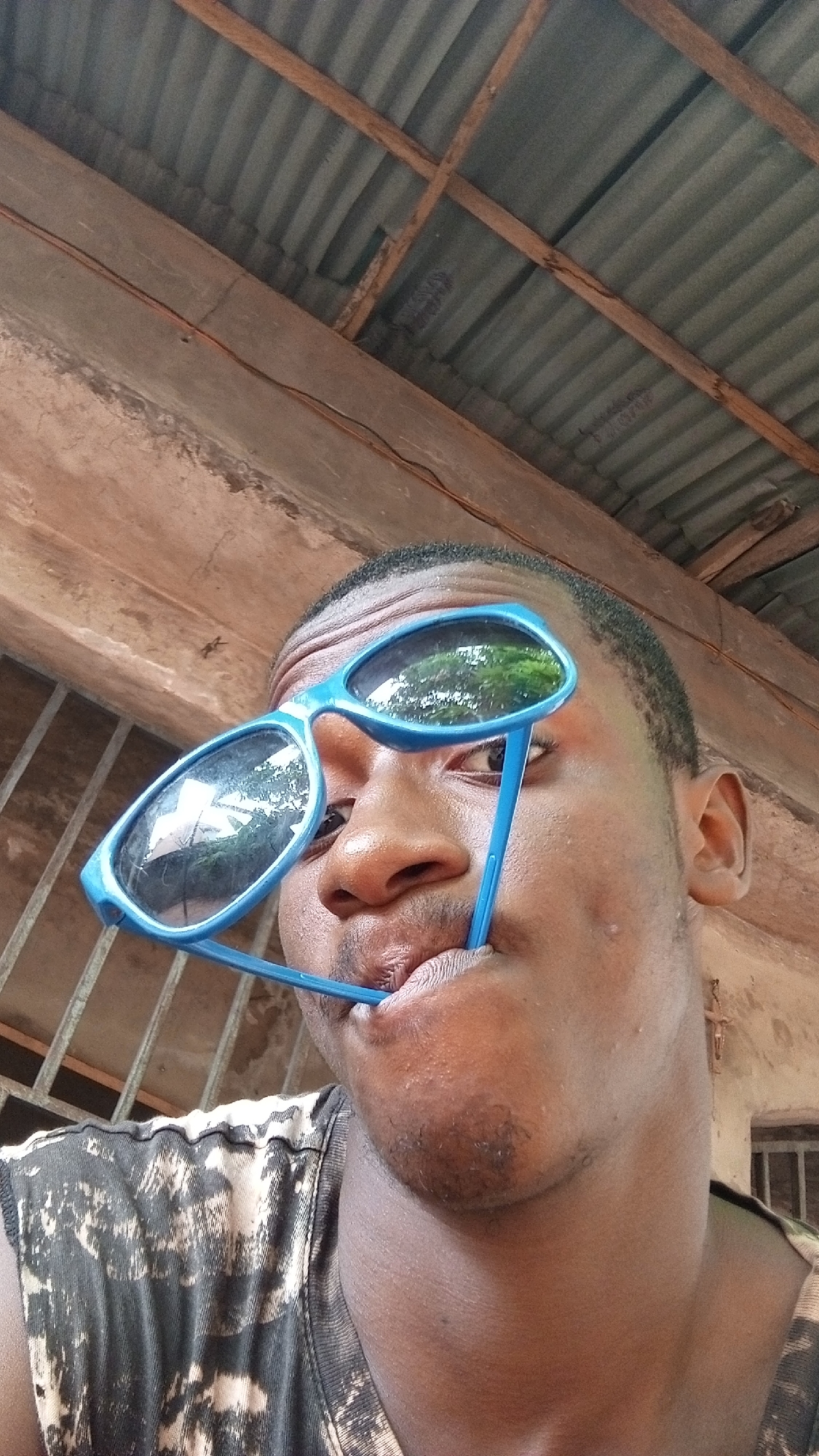 The image captures a close-up, upward-angled portrait of a young man, appearing to be of African descent. He has short dark hair and stubble on his chin. He is wearing a dark sleeveless shirt with a lighter, abstract or tie-dye pattern.
The man is engaging in a playful pose, holding a pair of blue-framed sunglasses in an unconventional manner. One lens of the sunglasses partially covers his left eye and forehead, while one of the arms of the frame is held between his lips and chin. The lenses of the sunglasses reflect green foliage, likely from trees or bushes, and a bright sky, indicating an outdoor or partially outdoor setting.
The background primarily features the underside of a corrugated metal roof, supported by visible wooden beams. Below this roof structure, there are textured, light-brown walls or concrete elements. In the lower right background, faint hints of a barred window or opening are visible, along with a small, cross-like decoration on a wall surface.
The lighting suggests it is daytime. The man's expression is neutral and calm, implying a casual or posed moment for the camera. There is no discernible text visible in the image. The overall setting appears to be a simple, possibly semi-sheltered outdoor area in what could be a residential or communal space.