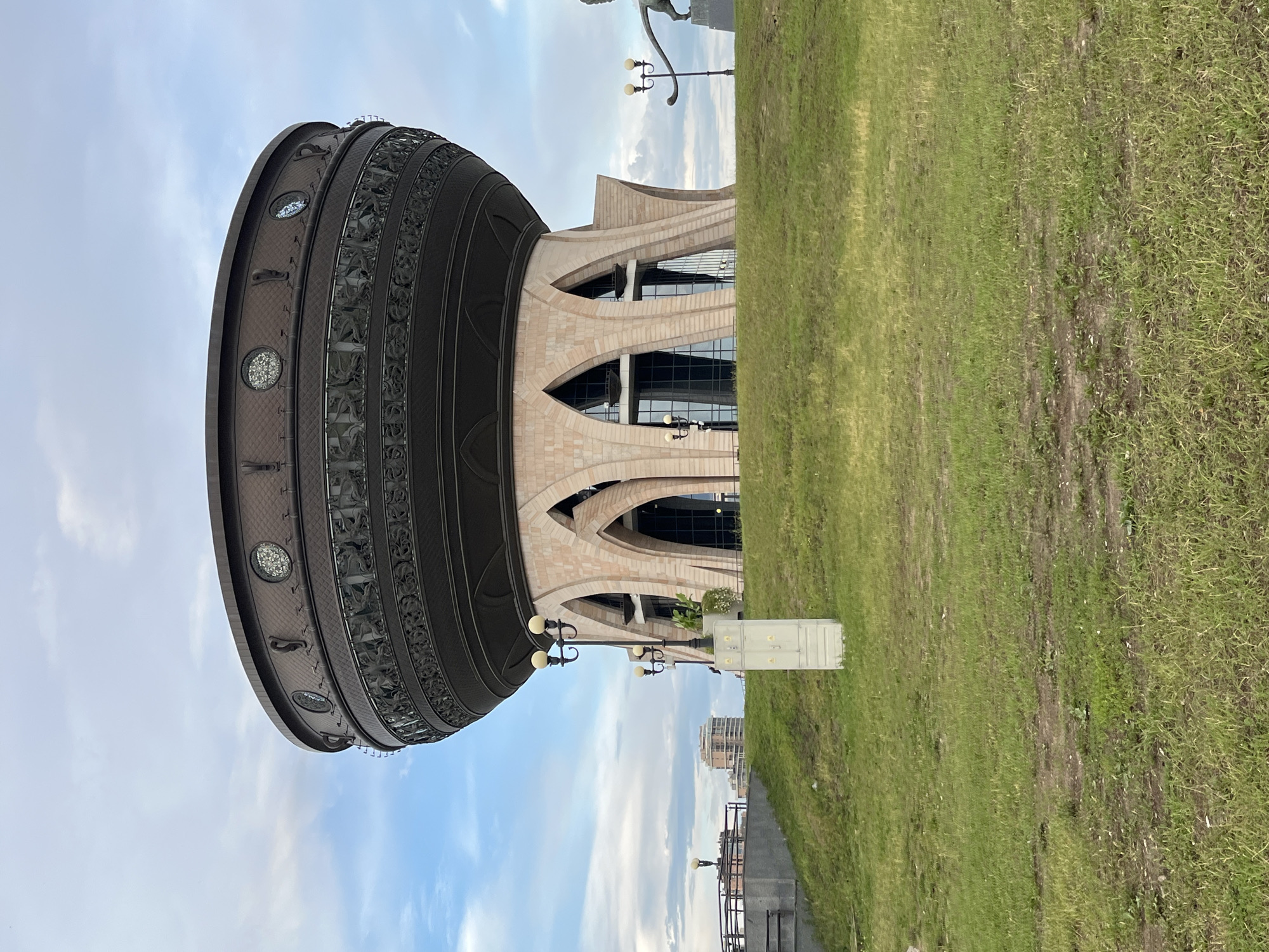 The image shows the Family Center "Kazan" in Kazan, Russia, also known as the "Chasha" or "Cup". The building is a large, cauldron-shaped structure with a dark bronze, ornate upper section featuring circular windows and decorative reliefs. The lower part of the building is made of light-colored stone with large arched openings. The building sits on a grassy hill. In the foreground, there is a stretch of green grass with some patches of bare earth. A light-colored electrical box is visible on the grassy slope, along with a lamppost with two globe-shaped lights. In the background, a lamppost with a single light is visible, along with the edge of a walkway or railing. Further in the distance, there are buildings and a statue of a winged lion. The sky is partly cloudy with a mix of blue sky and white clouds. The overall scene is an outdoor view of a distinctive architectural landmark.