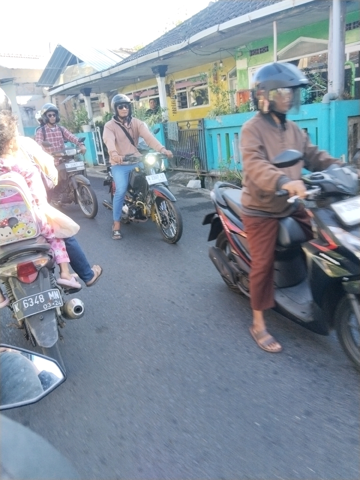 The image depicts a street scene in Banjararum, Indonesia, with several individuals on motorcycles. In the foreground, a person wearing a brown jacket and brown pants rides a dark-colored scooter. Their face is obscured by a helmet with a clear visor, and they are wearing sandals. To their left and slightly behind, another person on a motorcycle is visible. This individual wears a light brown hooded jacket and blue jeans, also with a helmet. Further to the left, a child is seated on the back of a motorcycle, wearing a backpack adorned with cartoon characters and pink pajamas. The child's legs are extended, and they are wearing pink slippers. The motorcycle in the foreground has a license plate that reads "K 6348 MN 03-24". The background shows residential buildings with colorful fences. The road surface appears to be asphalt. The lighting suggests it is daytime.