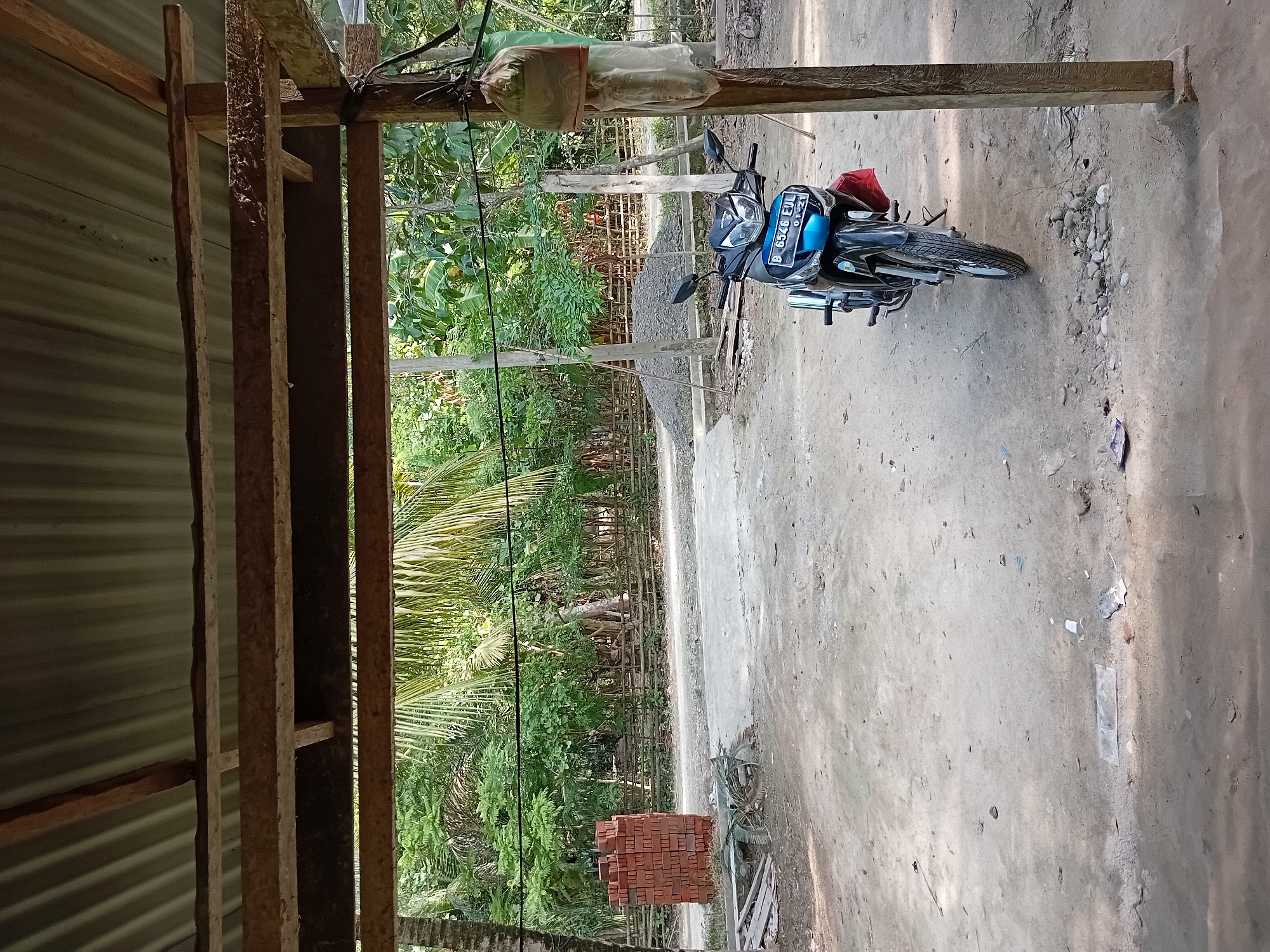 This is an outdoor photograph taken in Aluebuya, Indonesia. The foreground features a dirt ground surface with scattered debris. A motorcycle with a license plate reading "B 6546 EUL" is parked on the dirt. To the left, a stack of red bricks is visible, and to the right, a wooden pole supports two hanging bags. In the background, a lush green canopy of trees and foliage forms a natural backdrop. The scene is partially covered by a corrugated metal roof supported by wooden beams, suggesting a covered outdoor area or a simple structure.