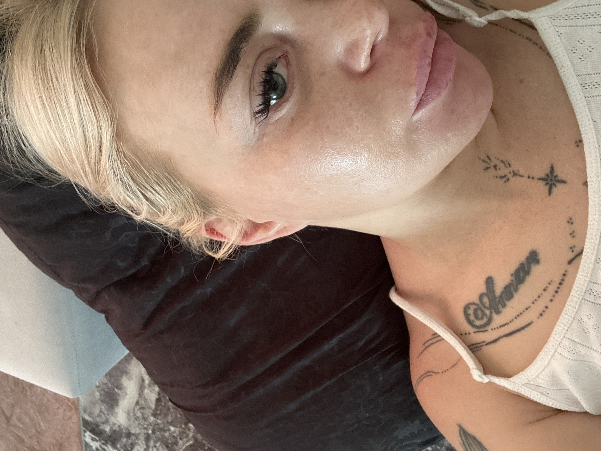 A close-up, over-the-shoulder shot shows a woman with blonde hair and fair skin, possibly in a home environment. She is lying down, her head resting on a dark, wrinkled surface. A white strap from her top is visible on her shoulder and chest, where there are several tattoos, including script and abstract designs. Her face is partially visible, with her eye open, looking towards the camera. The lighting highlights the texture of her skin. The background includes a blurred marble-like surface and a hint of a light blue surface. The scene is indoors, with no specific city or location identified.