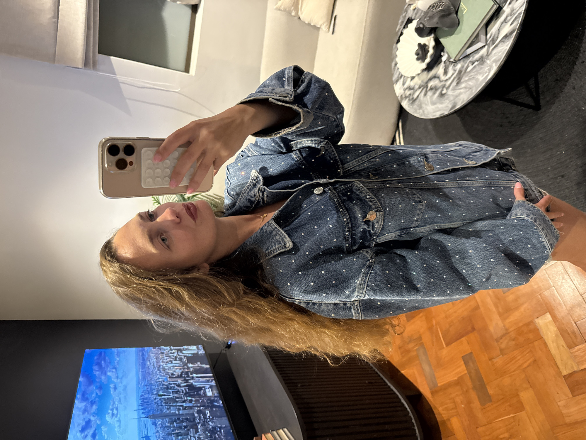 A woman is taking a selfie in an indoor setting. She is holding a smartphone in her right hand, and the camera is pointed at her face. She has long, blonde hair and is wearing a blue denim jacket with white polka dots. In the background, there is a TV displaying a cityscape with a blue sky. Adjacent to the TV, a wooden parquet floor is visible. In the distance, a sofa is present, along with a black and white table featuring books and decorative items. The exact location within Cape Town, South Africa, cannot be determined from the visual information provided.
