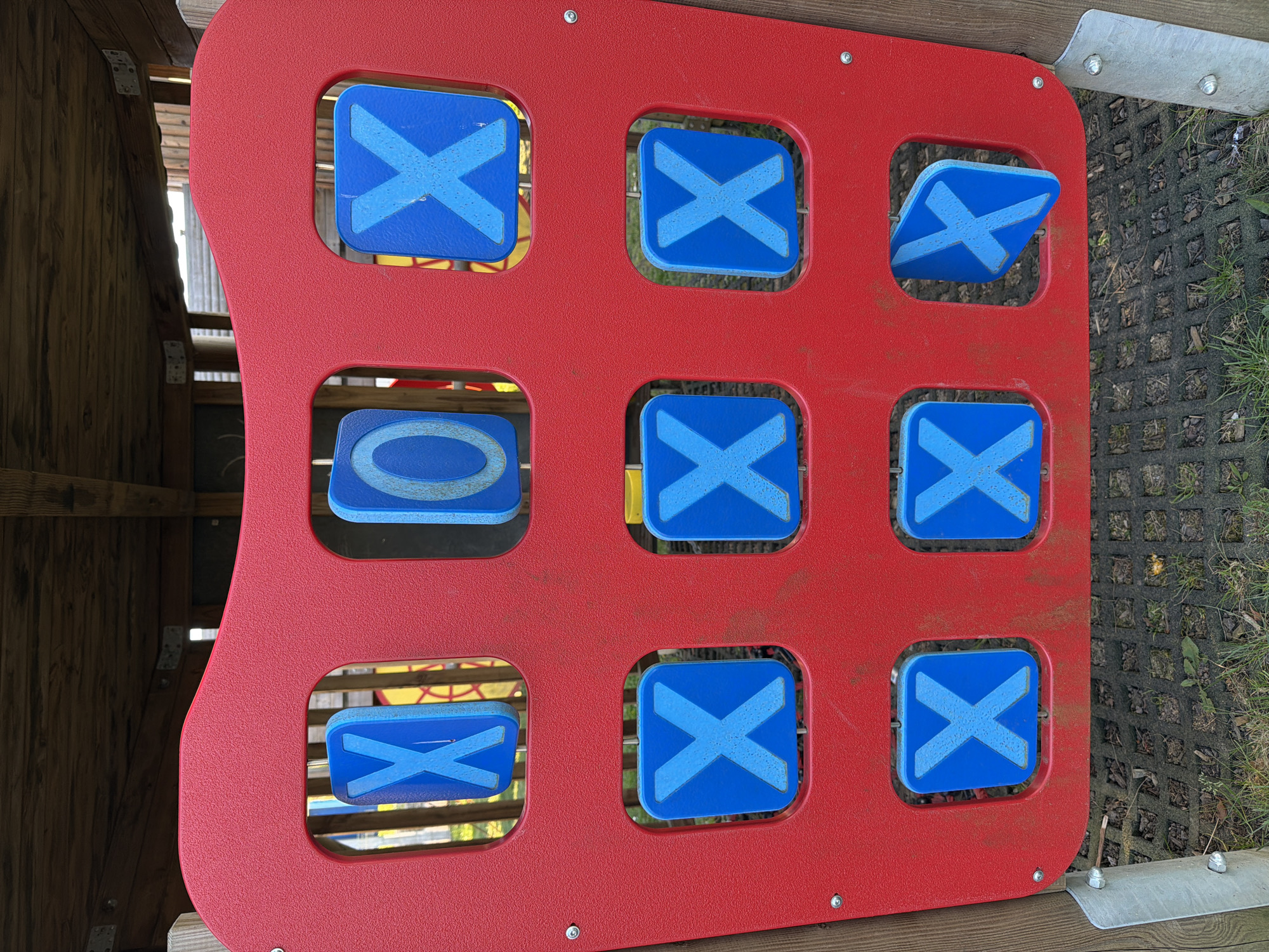 This image displays a red outdoor Tic-Tac-Toe (Noughts and Crosses) game panel, likely situated in a playground in Niederanven, Luxembourg. The panel is made of textured, red plastic, showing some signs of wear and dirt. It features nine square cutouts, each containing a rotatable blue plastic piece. Most of these pieces are currently displaying a light blue 'X' symbol. Specifically, eight 'X' symbols are visible across the board, arranged in various rows and columns. One blue piece in the middle-left position shows a light blue 'O' symbol. A piece in the top-right corner is partially turned, showing its edge, but still displays an 'X'.
The arrangement of 'X' symbols indicates multiple winning lines for the 'X' player, including horizontal wins in the top and bottom rows, vertical wins in the second and third columns, and diagonal wins. The game board is attached to a wooden structure on the left, possibly part of a playhouse or climbing frame. To the right, a dark grey grid-patterned ground surface, possibly rubber matting or concrete pavers, is visible with grass growing through. Hints of other yellow and blue playground equipment can be seen in the background through the panel's openings. The scene is illuminated by natural daylight, without strong shadows, suggesting an overcast day. No text is visible, only the game symbols.
