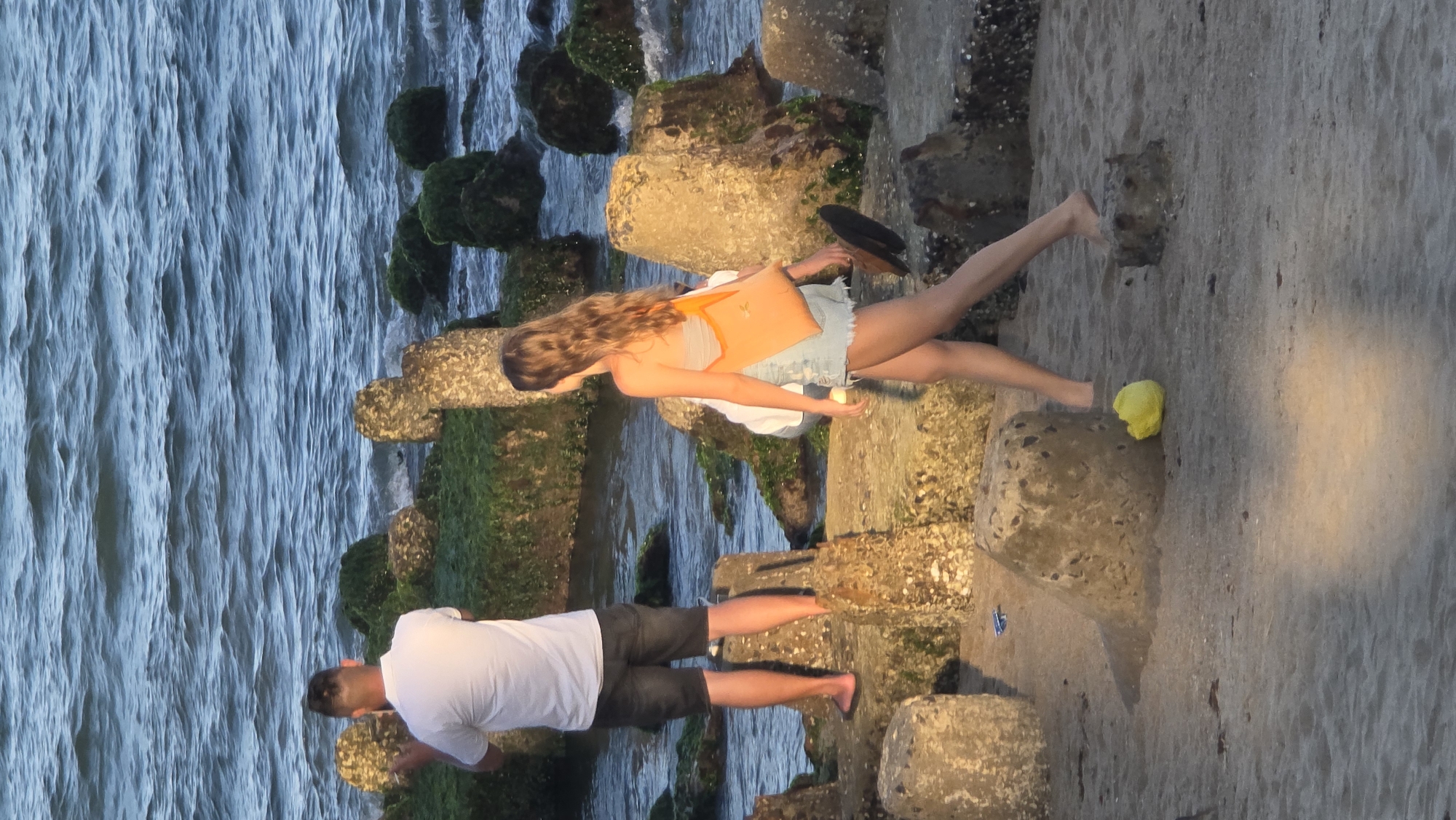 The image captures a serene coastal scene in Nha Trang Ward, Vietnam, likely during late afternoon or early evening, with warm sunlight casting a golden glow. Two people are seen walking barefoot along a rocky shoreline.
In the foreground and middle ground, large, irregular concrete blocks, many covered in green moss or algae, form a barrier or pathway separating the land from the water. To the left, a calm body of water, presumably the ocean, extends outwards with visible ripples on its surface. To the right of the rocks, a sandy or dirt path is visible.
A woman with long, wavy brown hair, wearing a light-colored top and denim shorts, walks on the rocks. She is carrying an orange shoulder bag and holding a dark hat in one hand. A small, crumpled yellow object rests near her left foot. Further along the rocks, a man in a white t-shirt and dark shorts is also walking, seemingly looking towards the water. The scene suggests a leisurely stroll by the sea. No text is visible.