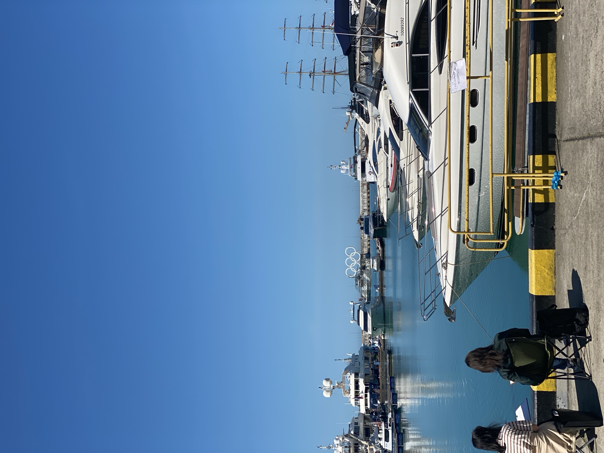 The image captures a vibrant marina scene under a clear blue sky. In the foreground, two individuals are seated on a dock, facing away from the camera. One person, with long dark hair and wearing a green jacket, sits in a green folding chair. To their left, another person with long dark hair and wearing a striped top sits on a lower chair or stool, seemingly engaged with papers or a book.
The dock features distinctive yellow and black striped safety markings. Beyond the dock, numerous boats and yachts are moored in the calm, blue-green water. These include several large, white motor yachts with sleek designs, some displaying prominent hull numbers like "10089392". One yacht has a white sign hanging from its railing, with visible Cyrillic text that reads "ПО ПОРУЧЕНИЮ ХОЗЯИНА" (BY ORDER OF THE OWNER).
Further down the marina, several older-style sailing ships with multiple tall masts are visible. In the distance, white Olympic rings, a clear indicator of the location, stand prominently against the sky. The bright sunlight suggests it is daytime, casting clear reflections on the water and strong shadows on the dock. The overall atmosphere conveys leisure and tranquility in a bustling port setting. Given the Olympic rings, the location is identifiable as Sochi, Russia.