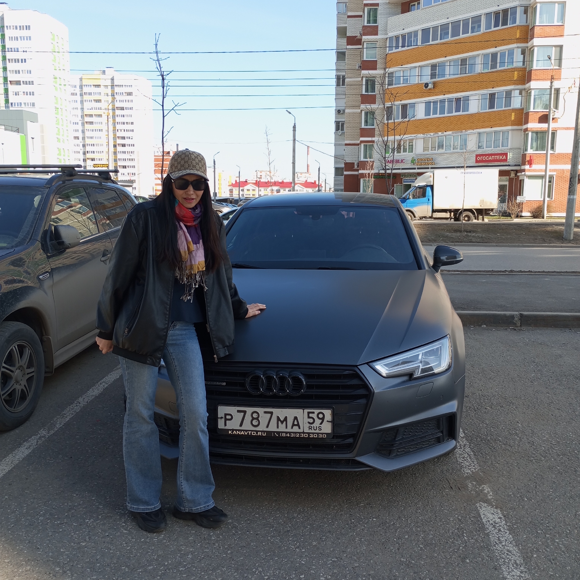 The image depicts a woman posing next to a dark grey matte Audi car in an urban setting, likely a parking area in Izhevsk, Russia. The scene is set on a clear, sunny day, with bright blue skies and distinct shadows.
The primary subject is a woman with long dark hair, wearing a patterned baseball cap (resembling a Gucci design), dark sunglasses, a black leather jacket, a multi-colored scarf, blue jeans, and black sneakers. She stands casually with her right hand resting on the hood of the Audi, which appears to be an A4 model.
Behind the woman and the Audi, another dark SUV is partially visible on the left. The background is dominated by several tall, multi-story residential buildings with various architectural details and color schemes, including white, green, orange, and brick. Numerous commercial signs are visible on the ground floors of these buildings, written in both English and Cyrillic script. These signs include "VAPESHOP," "FABERLIC," "ОГОСАПТЕКА" (pharmacy), "ЗОЛОТАЯ ТАБАКЕРКА" (tobacco shop), "ПРОДУКТЫ" (groceries), "ТУРАГЕНТСТВО" (travel agency), and "ГРУЗОПЕРЕВОЗКИ" (cargo transportation).
Further down the street, a white and blue delivery truck is parked. Power lines crisscross the upper part of the image, and a few bare tree branches are visible, suggesting either early spring or late autumn.
The Audi's front license plate is clearly legible as "P787MA 59 RUS," indicating registration in Perm Krai, Russia. Below the license plate, the text "KANAVTO.RU" and a phone number "(843)230 30 30" are displayed. The car's grille also features a partially visible "quattro" emblem. The overall atmosphere is casual, capturing a moment in a busy residential and commercial area.