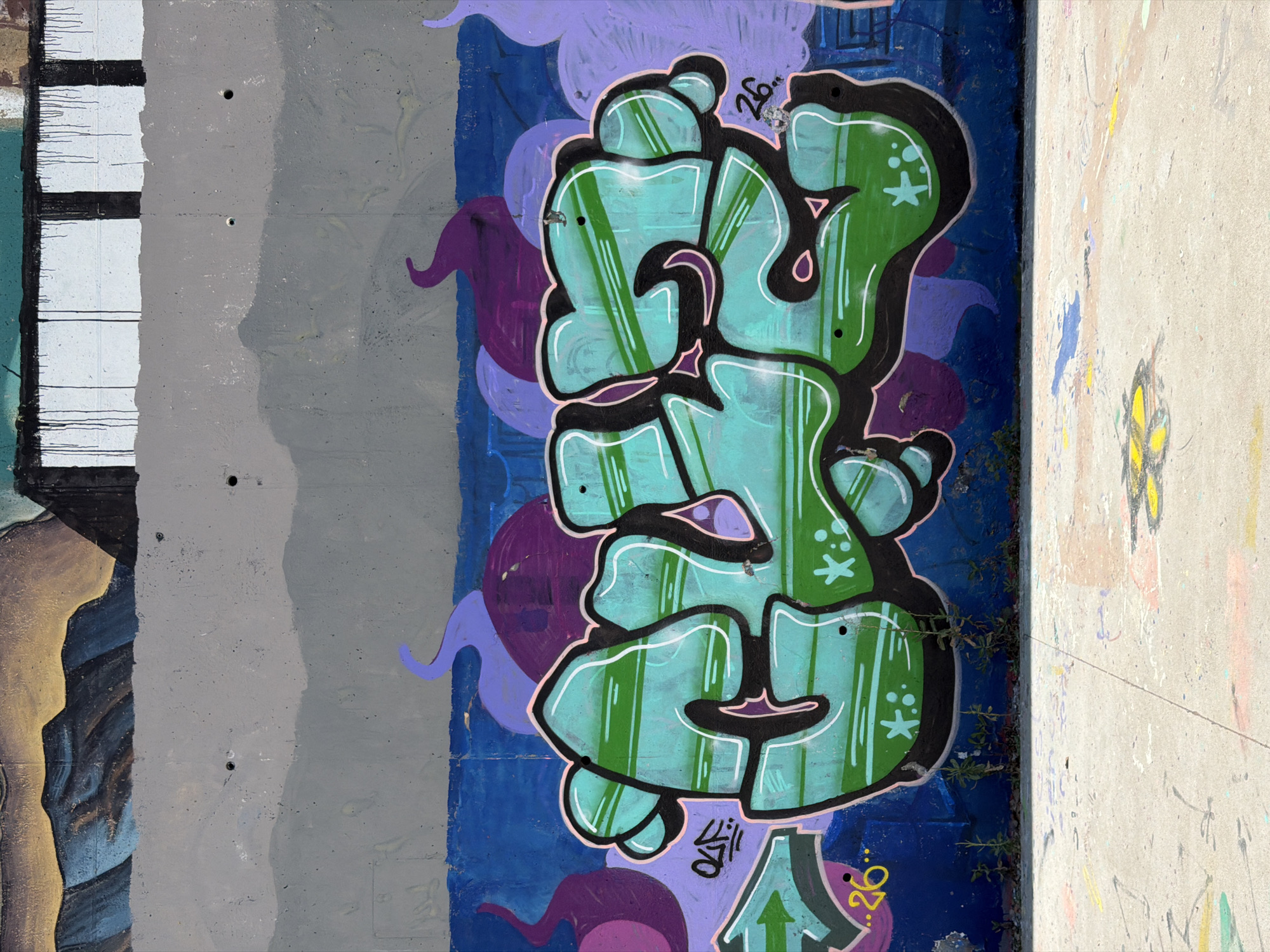The image shows a section of a concrete wall covered in vibrant graffiti art. Dominating the foreground is a large, stylized graffiti piece in shades of green and turquoise with thick black outlines. This lettering appears to be in a bubble style. Interspersed within the graffiti are smaller graphic elements like stars and dots, as well as what appears to be a tag or signature in black and purple. The background of the wall is a layered composition of colors, with a horizontal band of deep blue beneath the graffiti, above which is a large expanse of mottled gray concrete. Further up the wall, there's a painted section depicting abstract mountainous forms in blues and golden yellows. The bottom of the image shows a concrete ground surface, also marked with various colorful paint splatters and faint spray paint designs. The overall impression is of an outdoor urban environment, likely a public space such as a skate park or an area designated for street art, characteristic of locations in cities like Barcelona, Spain, known for its active street art culture. There are no people or animals visible in the image.