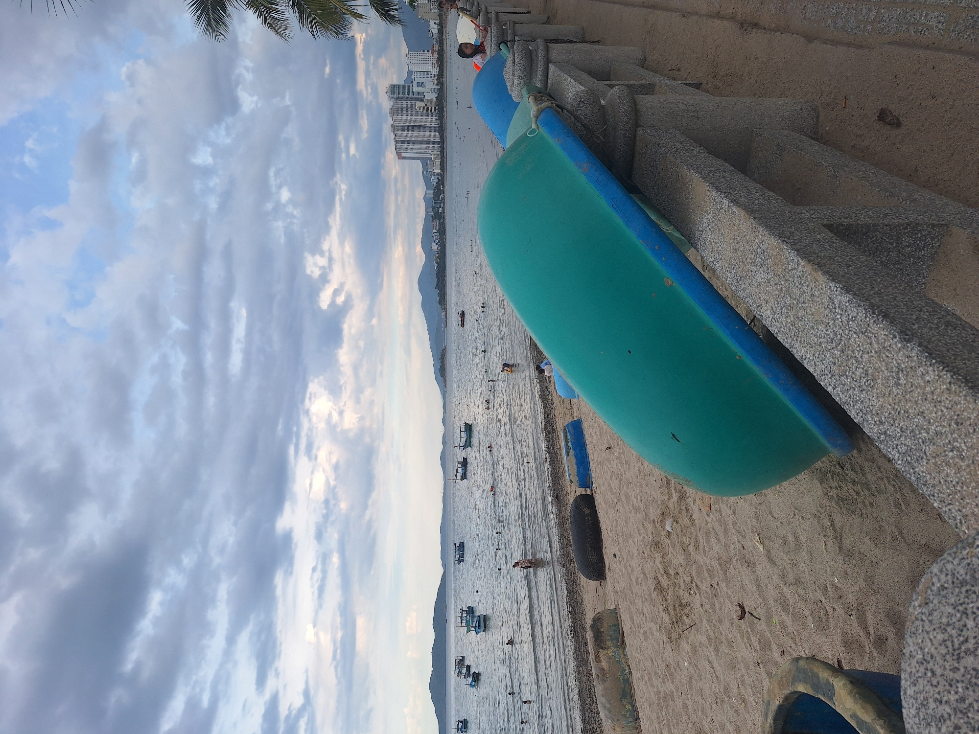 This is a photograph of the beach in Nha Trang, Vietnam. The scene is captured from a low angle, looking towards the sea. In the foreground, there are several overturned boats made of fiberglass, painted in green and blue, resting on the sandy beach. A stone railing with decorative pillars runs along the right side of the image, separating the beach from a paved walkway.
The sea stretches out in the background, dotted with numerous small fishing boats. People are visible in the water, swimming and playing. Further in the distance, a shoreline is lined with tall buildings, some of which appear to be hotels or apartment complexes. The sky is partly cloudy with a mix of grey and white clouds, suggesting it might be late afternoon or early evening. A few palm fronds are visible on the far right, indicating the tropical setting. The overall impression is one of a busy beach resort town.