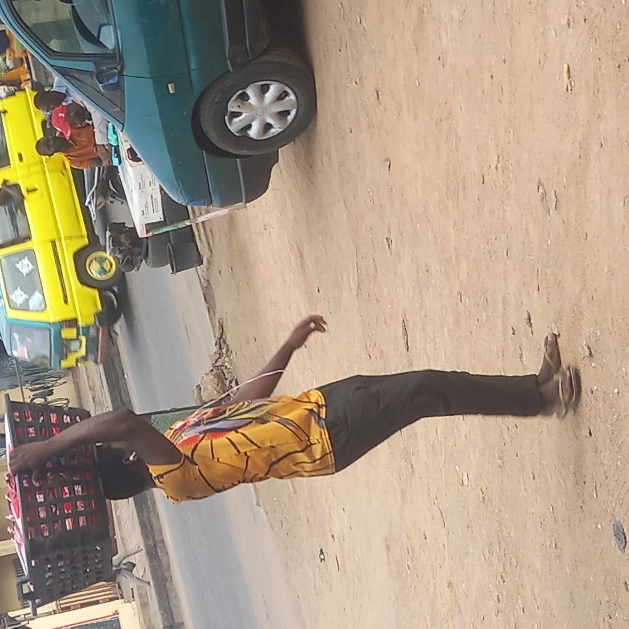 A man walks along a dirt and paved road in Dadi Riba, Nigeria. He carries a full plastic crate on his head. The crate appears to contain many red and white rectangular items. The man is wearing black pants, a yellow shirt with red and black patterns, and sandals. The road appears to be lined with vehicles, including a green car, a yellow van, and a blue tricycle. The photo is taken from an elevated perspective.