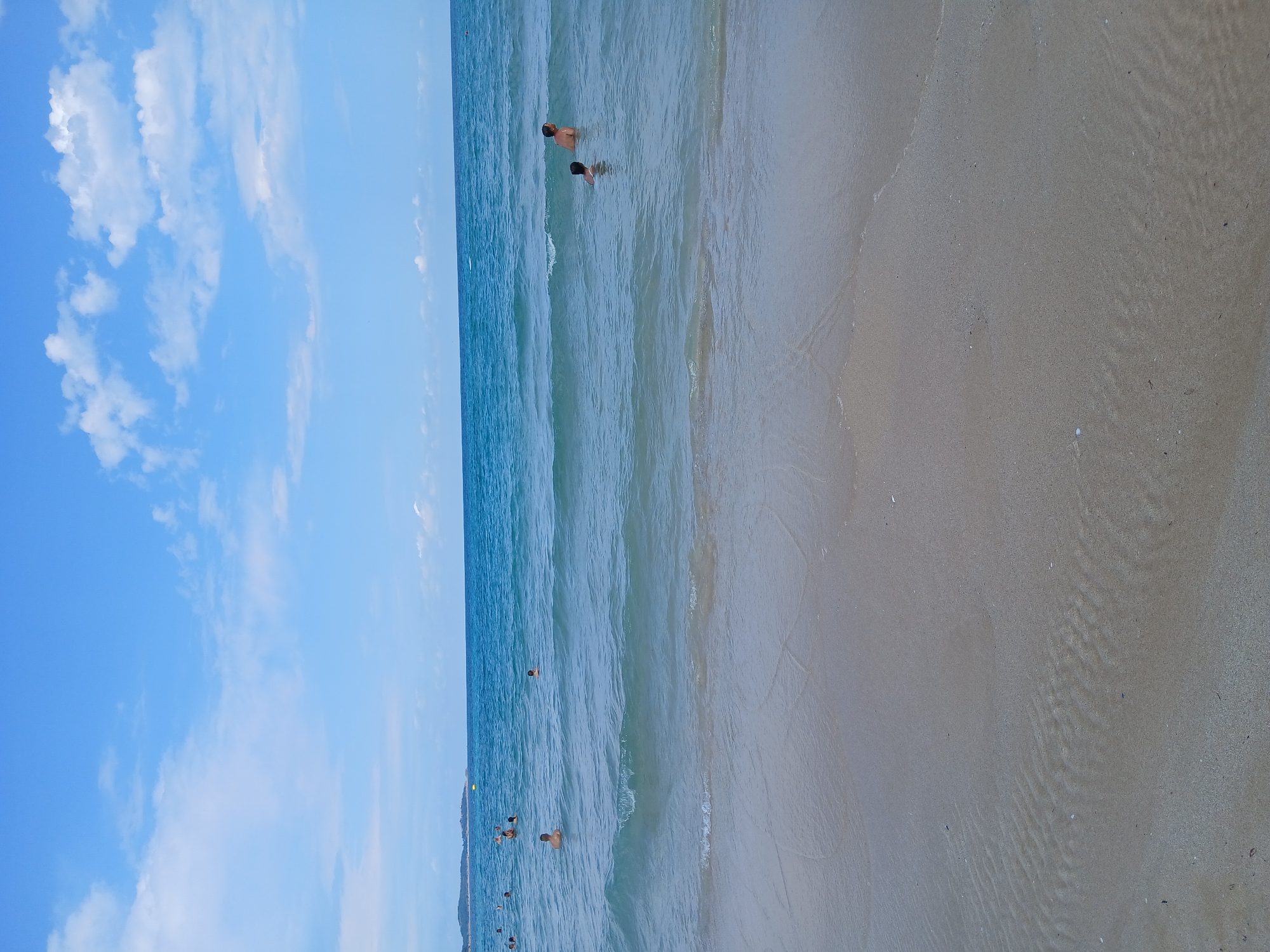 The image depicts a scene at a beach in Primorsko, Bulgaria. The sky is a clear blue with scattered white clouds. The sea is a vibrant turquoise, with gentle waves approaching the shore. Several people are visible in the water, swimming and enjoying the sea. The foreground of the image is occupied by a sandy beach, with subtle patterns left by the receding tide. In the distance, a coastline with some vegetation and structures can be seen.