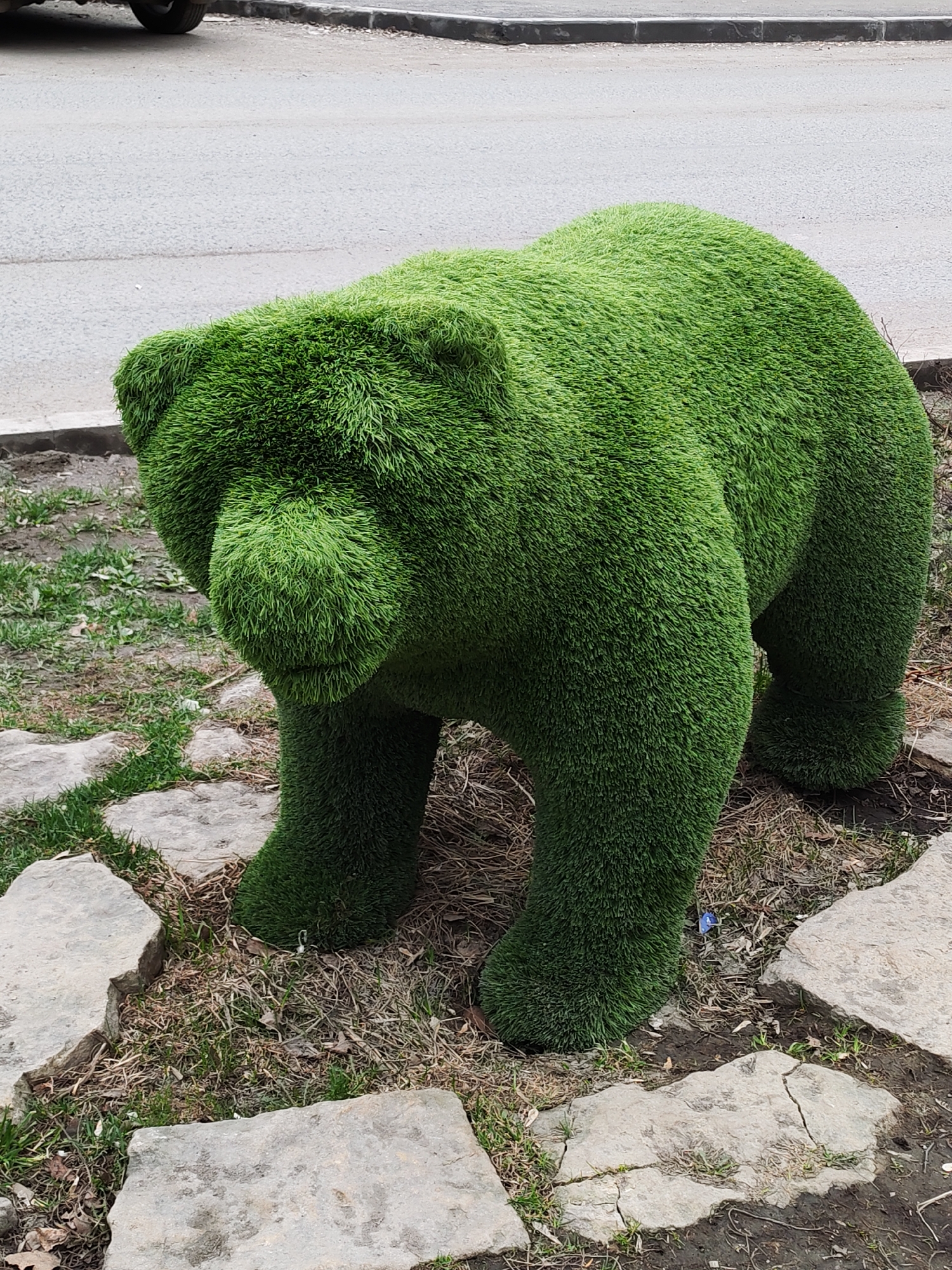 A full, medium shot captures a large, green, artificial grass bear sculpture. The sculpture is placed on a ground of dirt, stone slabs, and patchy grass. A street is partially visible in the background. The location is most likely in the city of Izhevsk, Russia.
