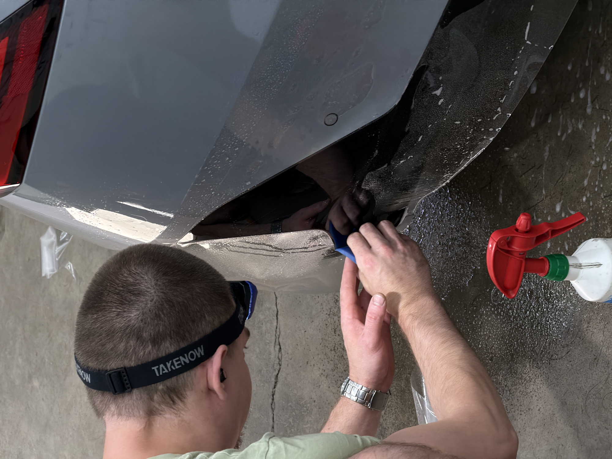 A person, wearing a light green t-shirt and a black headband with the word "TAKENOW" written on it in white letters, is applying a clear protective film to the rear bumper of a dark gray car. The person is leaning over the car, focused on the task. Their hands are visible, one holding a blue squeegee-like tool to smooth the film, and the other appearing to guide the film. The car's surface is wet, with water droplets and some suds visible, suggesting a cleaning or application process. A red-handled spray bottle with a white base is on the ground in the foreground, also indicating some form of liquid application. The background shows a concrete floor with some cracks and debris. The overall scene appears to be an automotive detailing or protection service. There is no indication of the specific location within Krasnoyarsk, Russia.