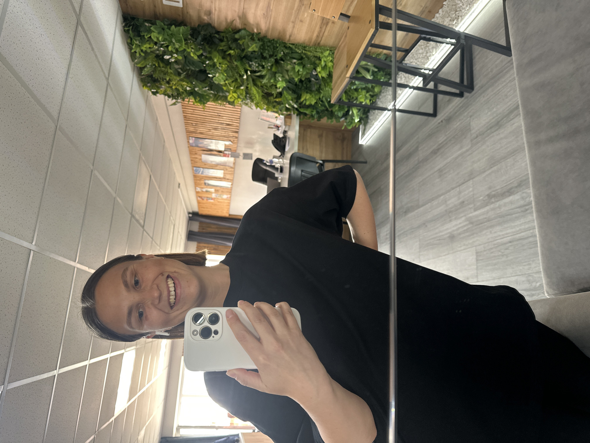 A woman smiles and takes a selfie in a commercial space. The background includes a ceiling with square panels, wood accents, and greenery on the wall. Modern tables and chairs are visible in the mirror reflection. The location is likely in Chaikovsky, Russia.