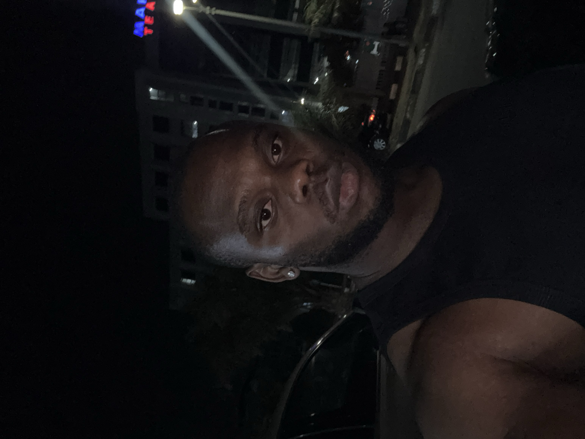 The media file presents a dark, portrait-oriented selfie taken at night, rotated 90 degrees counter-clockwise. The central subject is a dark-skinned man with a short, trimmed beard, looking directly into the camera. He wears a dark, possibly black, sleeveless top or tank top, revealing his muscular build. A small, shiny stud earring is visible in his left ear. His expression is neutral, with a slight pout on his lips.
The background is dimly lit, typical of a nighttime urban environment, and appears to be in Lugbe, Nigeria. Behind the man, a multi-story building is visible, illuminated by scattered artificial lights from windows and what seems to be a streetlight or lamppost. On the upper section of the building, a partial neon sign is discernible, displaying "MA" in blue letters and "TE A" in red letters. Faint outlines of trees and other distant light sources suggest an outdoor street scene or commercial area. A blurry shape in the distance to the right might be a vehicle. The overall lighting casts the scene in deep shadows with highlights on the man's face and parts of the background.