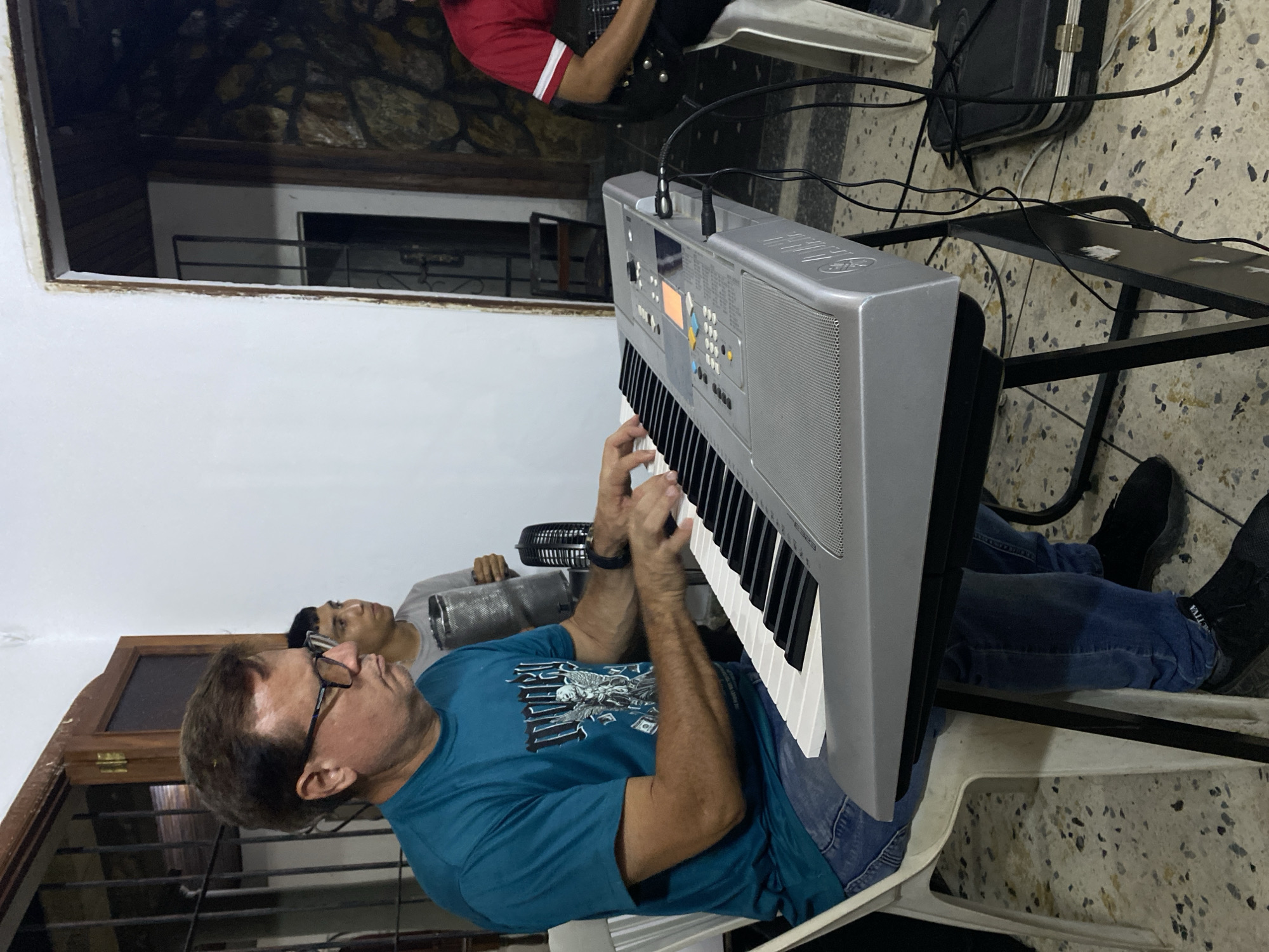In Tinaquillo, Venezuela, an indoor scene depicts three individuals engaged in musical activity. In the foreground, a male of apparent middle age, wearing glasses, a teal t-shirt with a graphic design, and blue jeans, is seated on a white plastic chair, actively playing a grey Yamaha electronic keyboard. His hands are positioned over the white and black keys. A black watch is visible on his left wrist. The keyboard rests on a black stand, with a black audio cable connected to its right side, and additional black cables are present on the floor.
Behind and to the right of the keyboard player, a younger male in a grey t-shirt holds a metallic, cylindrical percussion instrument. To the far right, partially visible, another individual in a red t-shirt with white stripes is seated, holding a black electric guitar. A black microphone with a mesh top is positioned near the keyboard. The floor is covered with light-colored speckled tiles. The background features a white painted wall to the left with a wooden doorframe revealing metal security bars, and a dark stone-textured wall to the right within another doorway or archway. A black hardshell case with metallic clasps lies on the floor to the right of the keyboard stand.