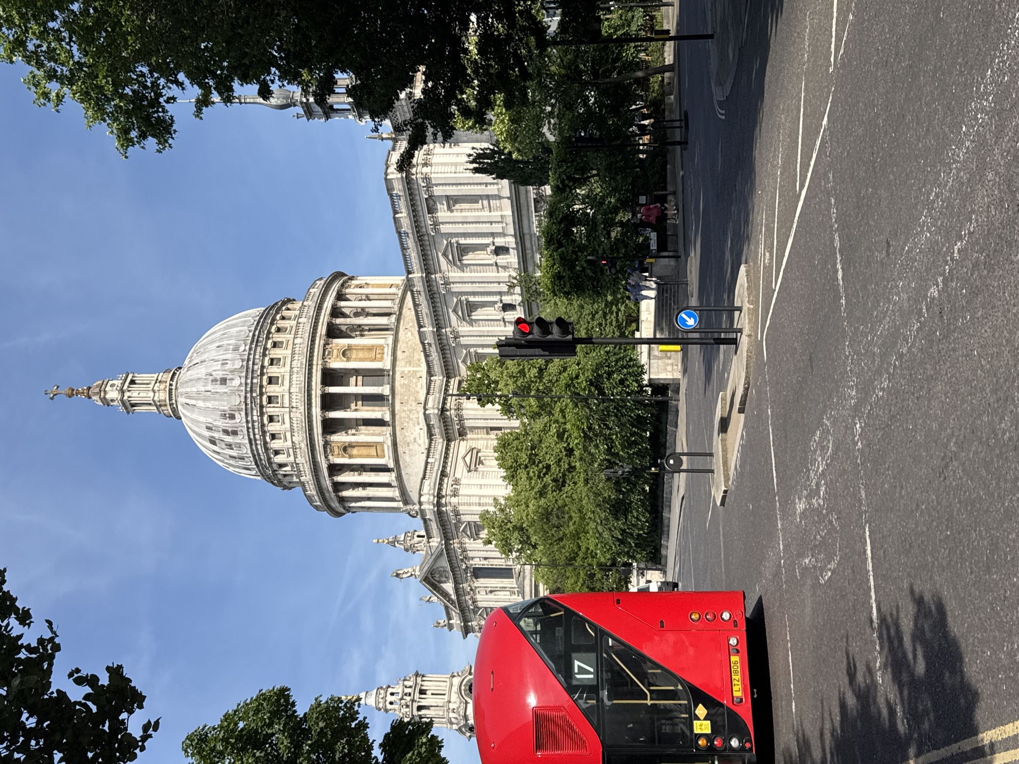 St. Paul's Cathedral, a well-known building in London, is seen from street level. The street is lined with trees, and there is a red double-decker bus in the foreground. Traffic lights are visible, showing red. There is a blue directional sign. The sky is clear and blue. There are people sitting on a bench. The bus is marked with route number 17. The bus registration number is LTZ1806.