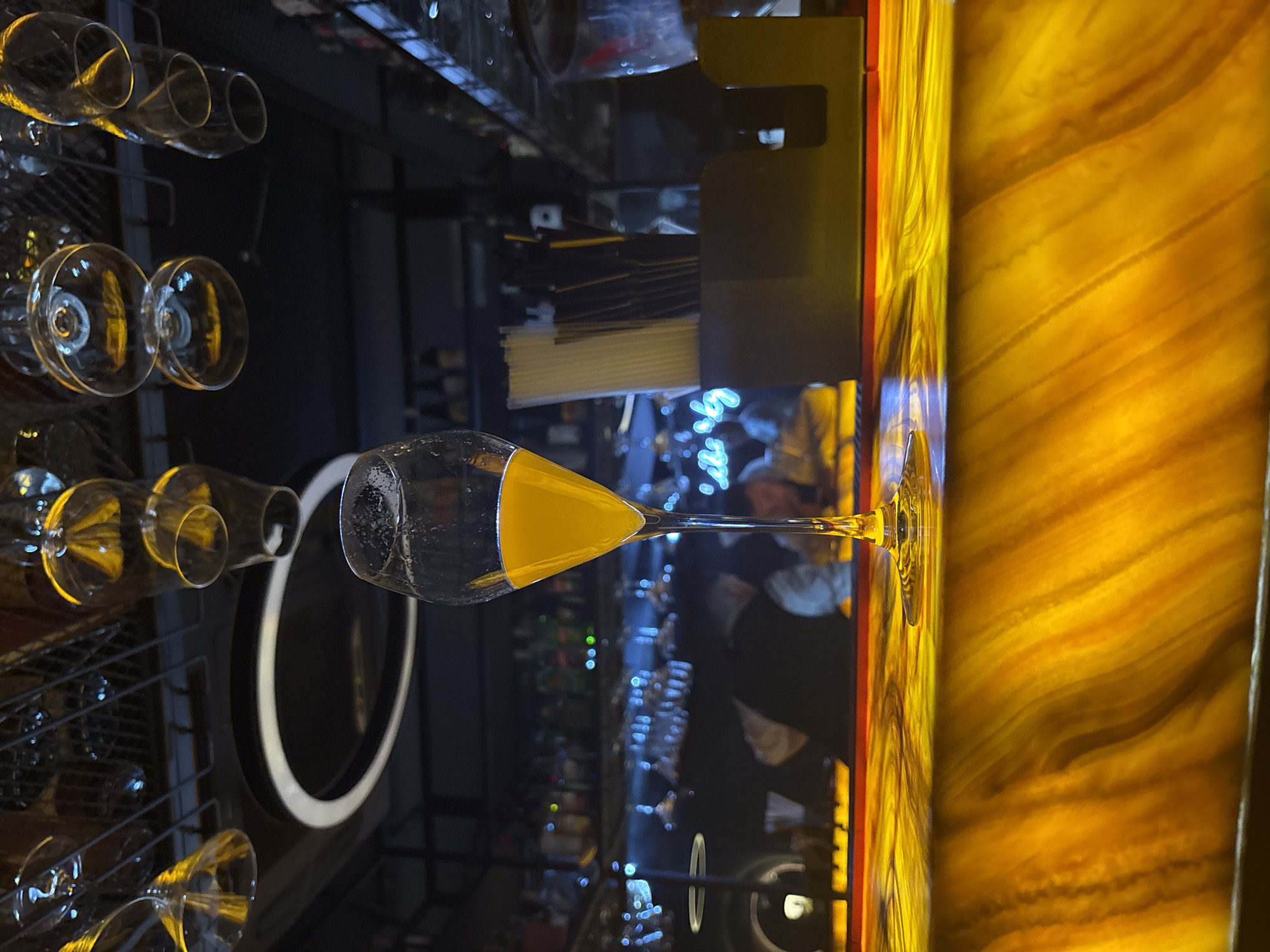 Here's a detailed description of the image:
The image shows a bar setting, possibly in a restaurant or lounge. The scene is brightly lit with warm, yellow lighting emanating from a textured wall or counter surface. The lighting creates a vibrant and somewhat abstract backdrop.
In the foreground, there is a wine or cocktail glass filled with an amber-colored liquid (likely a cocktail or wine). The glass is positioned as if it has just been served or is awaiting service.
Behind the glass, there is a rack or shelf holding multiple wine glasses. The glasses vary in size and shape, suggesting a range of beverage options. Other details visible in the background indicate a bar environment, like bar equipment and general lighting. There are dark tones that give a sense of depth and contrast, typical of an active nighttime establishment.
The image is vertically oriented.