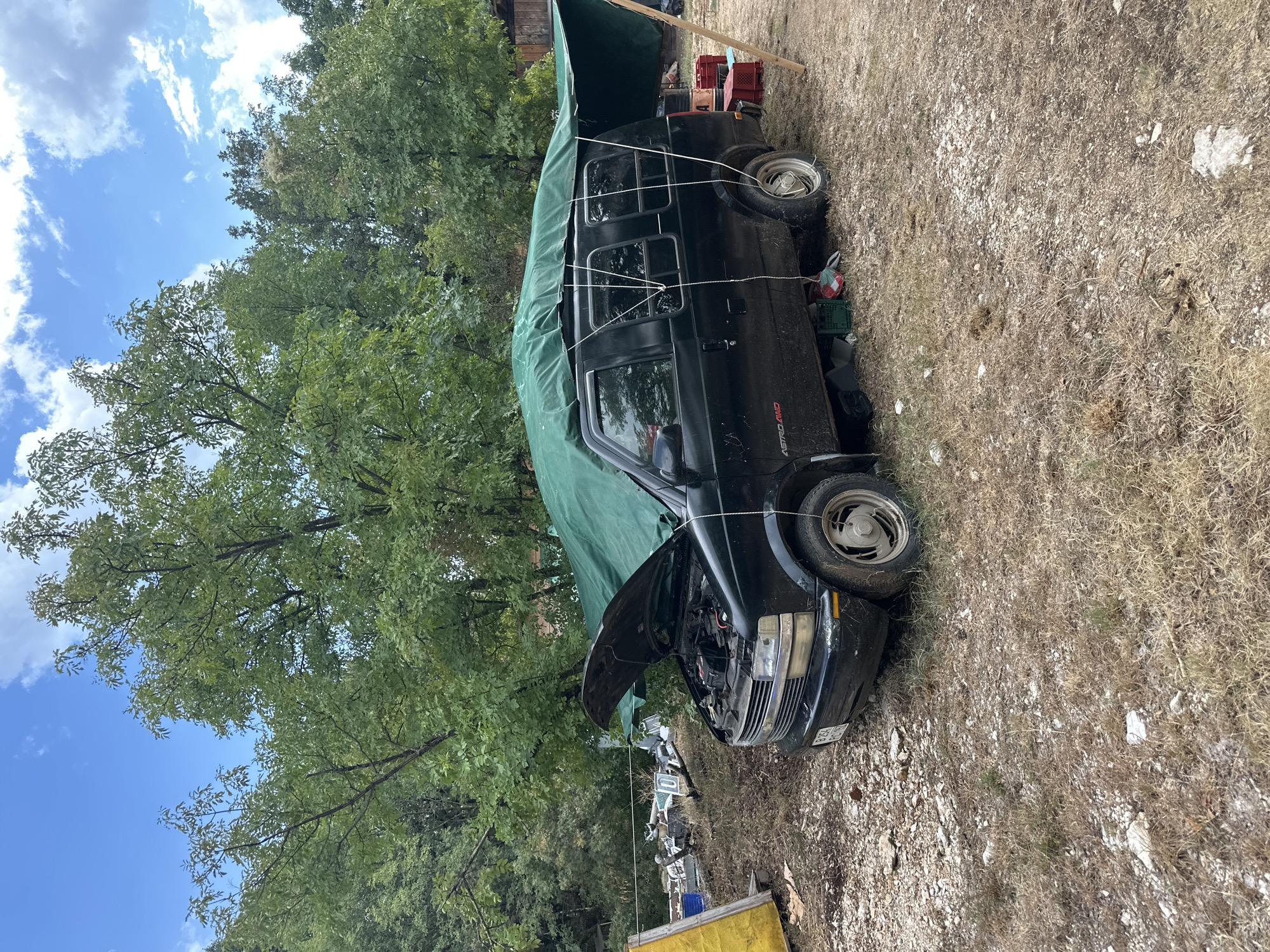 The image shows a black van with its hood open, covered by a large green tarp tied down with ropes. The van is parked on a gravelly area with dry grass and some scattered rocks. Trees with green leaves form the background, and a blue sky with scattered clouds is visible above. To the left of the van, there is a yellow tarp and some discarded items, including what appears to be old metal and plastic pieces. Red crates are stacked near the rear of the van, and a wooden pole is propped up against the tarp. The scene appears to be an outdoor, possibly rural or semi-abandoned location. There are no people visible in the image. The van has "ASTRO AWD" written on its side. The license plate is partially visible.