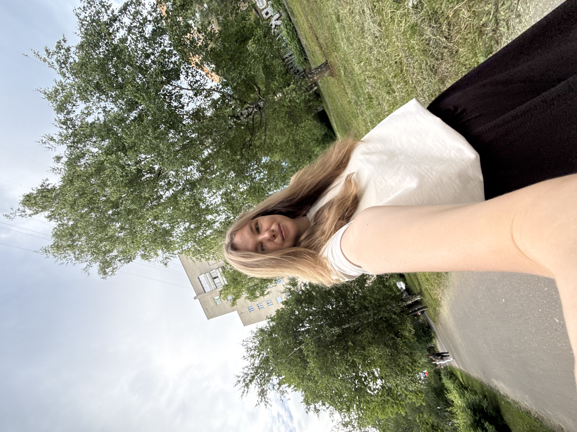 A blonde woman is taking a selfie outdoors. She is wearing a white t-shirt and a black skirt. She is standing in front of a building and trees. The image appears to be taken on a cloudy day. The location is in the city of Vladimir, Russia, near the intersection of Mira Street and Pionerskaya Street.