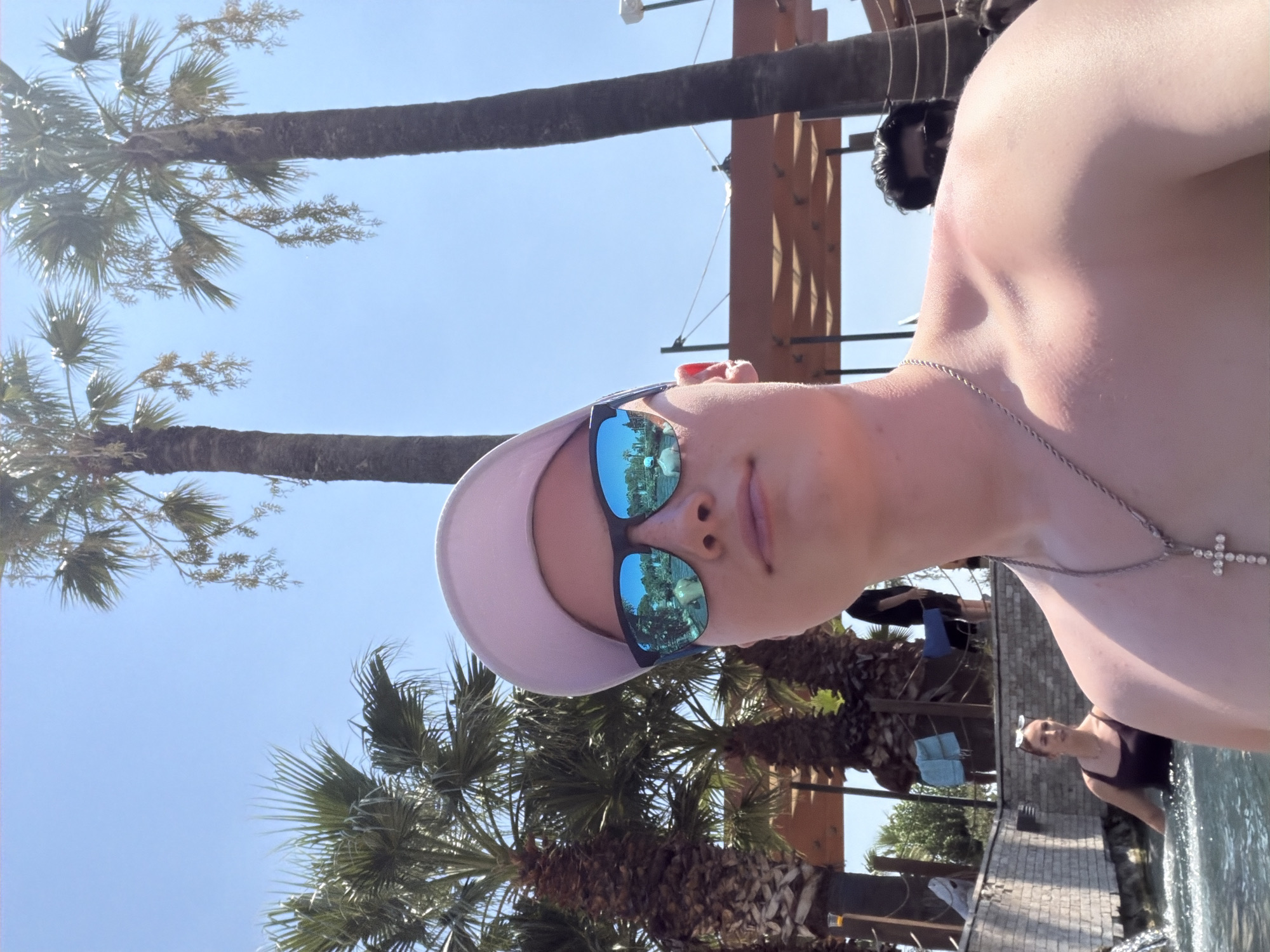 In Pamukkale, Turkey, on a sunny day, a young person in a light-colored baseball cap and sunglasses is taking a selfie in a pool with palm trees and a blue sky in the background. The person is wearing a silver chain with a cross pendant. In the background, a woman in a dark swimsuit is in the pool, and other people are visible. The setting appears to be a resort or a water park, with lounge chairs and an outdoor seating area.
