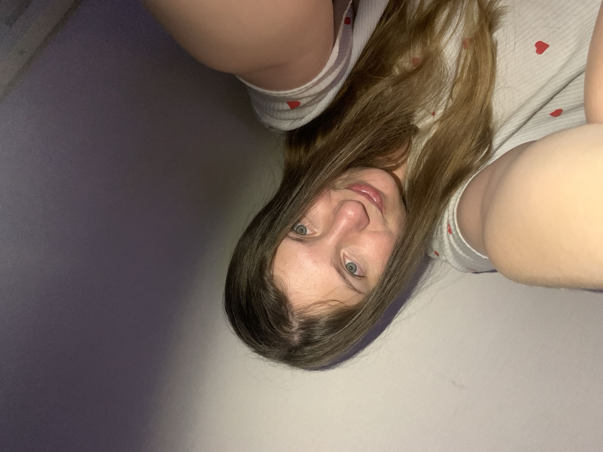 A close-up, rotated portrait image shows a young woman with long, medium-brown hair. Her blue-green eyes gaze directly at the camera, and she has a subtle, closed-mouth smile. She wears a light-colored top or garment adorned with small red heart patterns. Her shoulders and upper arms are visible on the periphery of the frame. The image appears to be taken from an overhead perspective, creating a slightly distorted view, and is oriented 90 degrees clockwise from an upright position. The background is an indistinct indoor setting, with a plain wall or surface that is darker grey-purple on the left and lighter off-white on the right, suggesting varied lighting. The overall scene conveys a casual, intimate moment indoors in Chaikovsky, Russia. No text is visible.