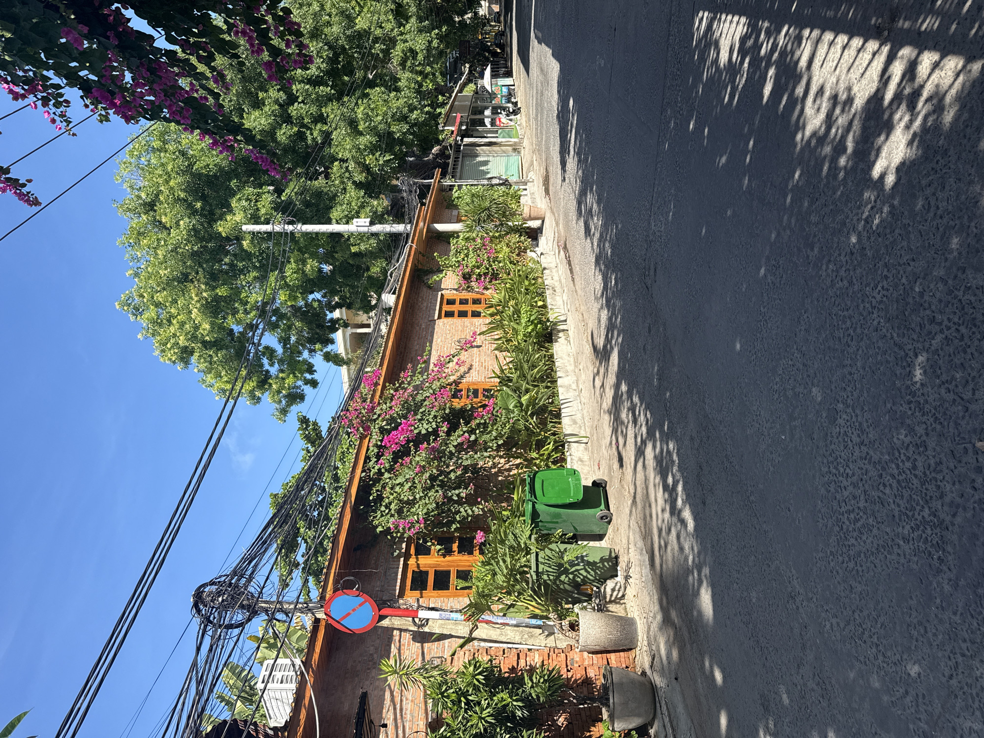 The image depicts a street scene in Nha Trang Ward, Vietnam. A brick building with orange-framed windows stands along the left side, partially covered by bougainvillea flowers. A green trash bin is placed on the sidewalk in front of the building. Power lines are visible above, with a "No Parking" sign attached to a pole. A paved road stretches into the distance on the right, dappled with shadows. Trees and other greenery are present in the background. The sky is clear and blue.