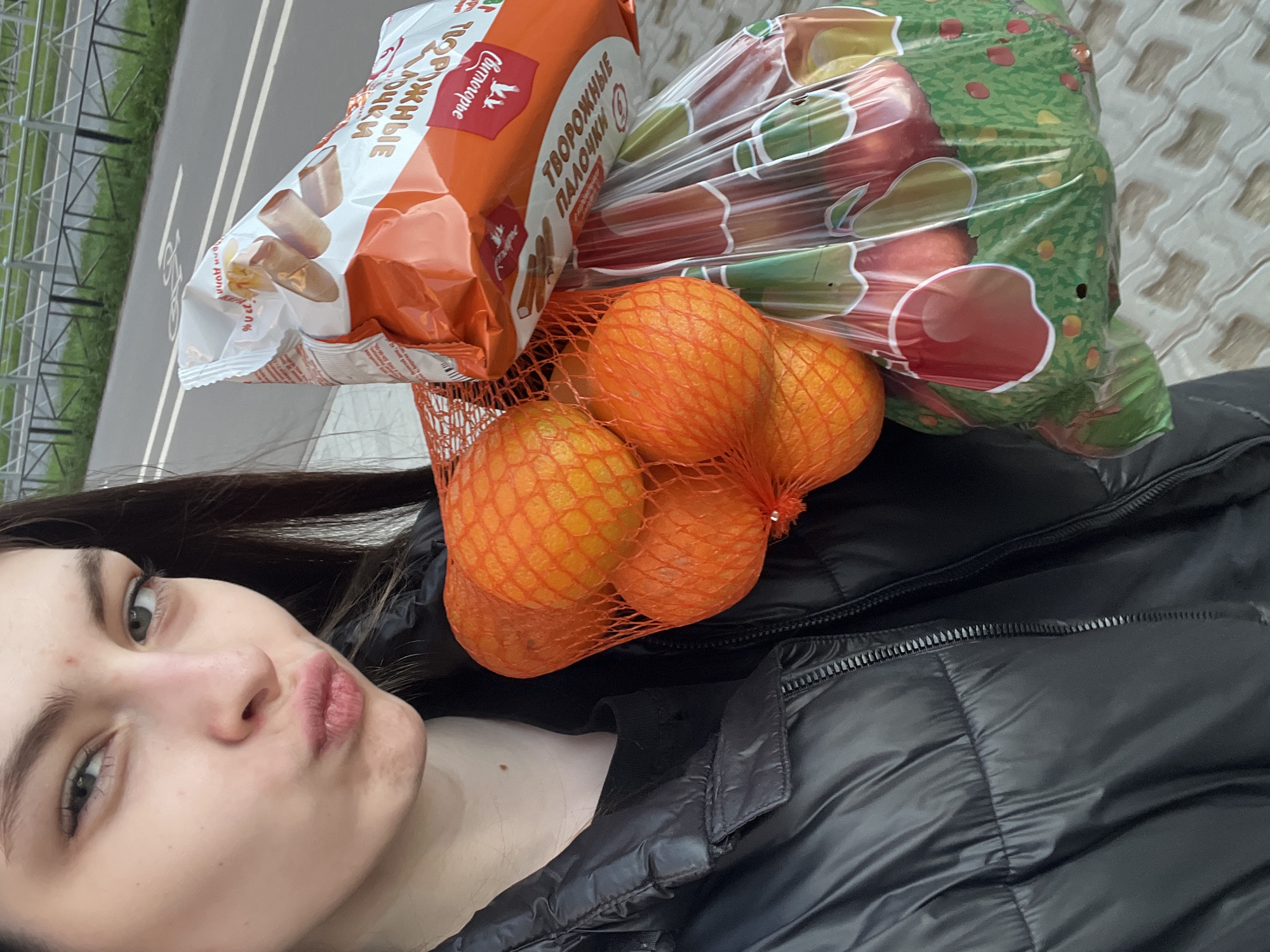 A young woman is holding groceries in front of her while outdoors. The groceries include a package of "Творожные палочки" (sweet cheese curd sticks), a bag of apples, and a net of oranges. The woman is wearing a black puffer jacket. A street with painted lanes is visible in the background. The location is likely Saint Petersburg, Russia, based on the Russian packaging of the "Творожные палочки."