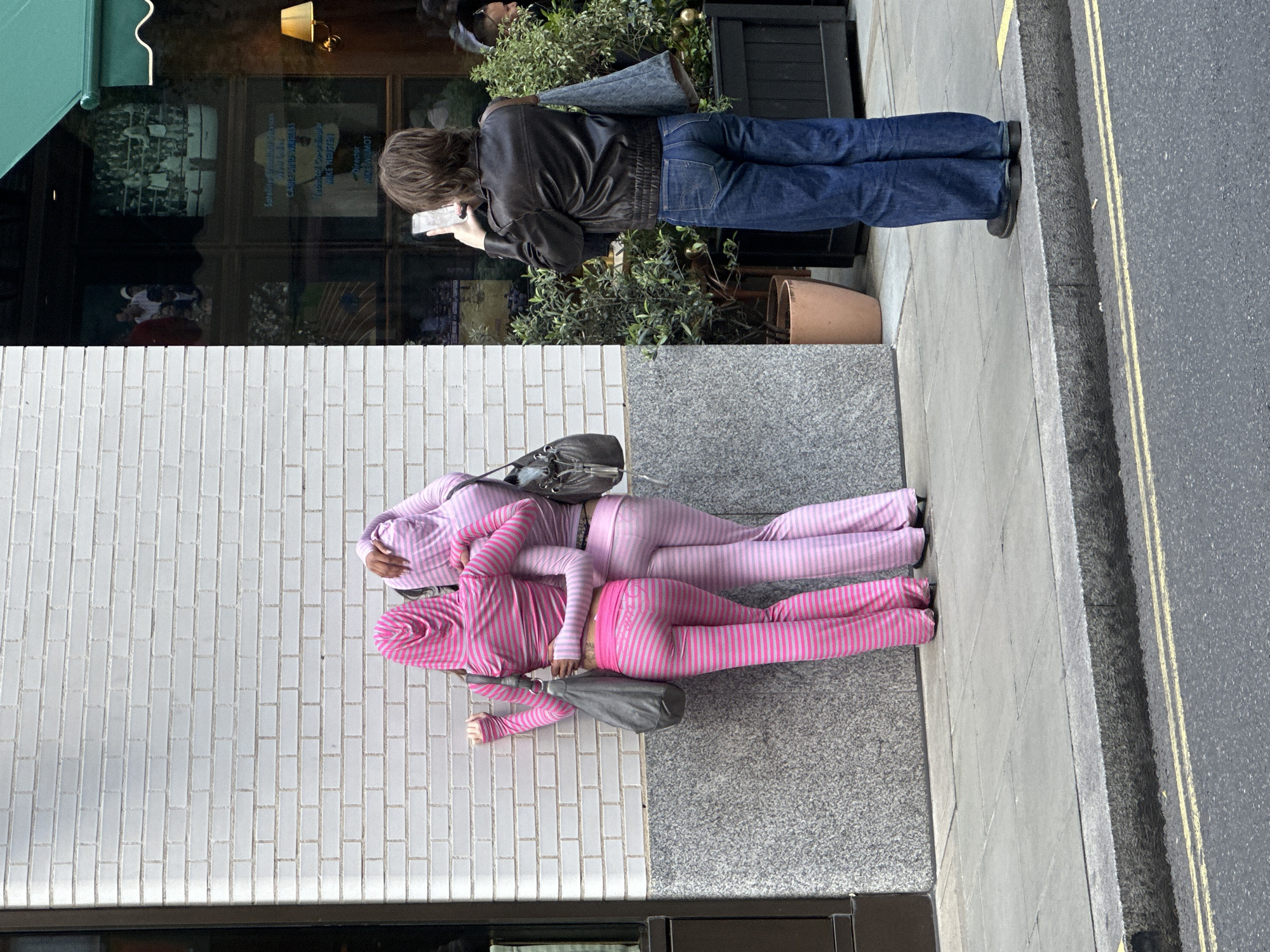 On a city sidewalk, two individuals wearing matching striped pink and lavender hooded tracksuits lay prone next to a building with white brick walls. Their bodies are parallel and close together, each with a grey shoulder bag. Adjacent to them, a person with long brown hair, wearing a black leather jacket and blue jeans, stands on the sidewalk, looking at a mobile phone. This person is positioned near a building with a green awning, behind which is a window reflecting the interior. The sidewalk edge borders a roadway marked with double yellow lines. The setting is urban, likely in London, United Kingdom.