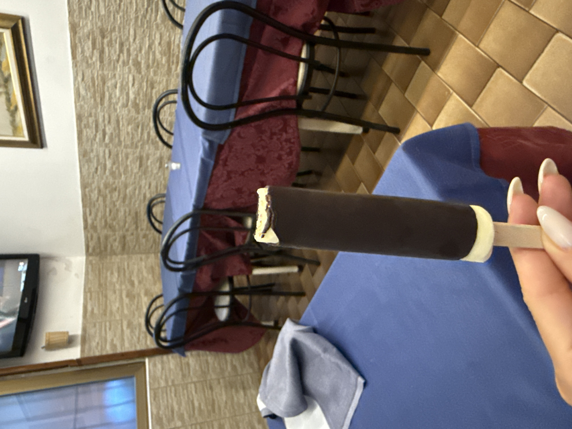 In a dimly lit indoor space, possibly a restaurant or dining area in Sesto San Giovanni, Italy, a person's hand with manicured fingernails holds a chocolate-covered ice cream bar. The ice cream bar has a dark chocolate coating over a lighter interior and is attached to a wooden stick. The background reveals several round tables covered with blue cloths and red tablecloths underneath. Black metal chairs with curved backs are arranged around the tables. The walls are tiled with a textured, light brown material. A television is mounted on the wall in the upper left corner. The floor is tiled with square, light brown tiles.