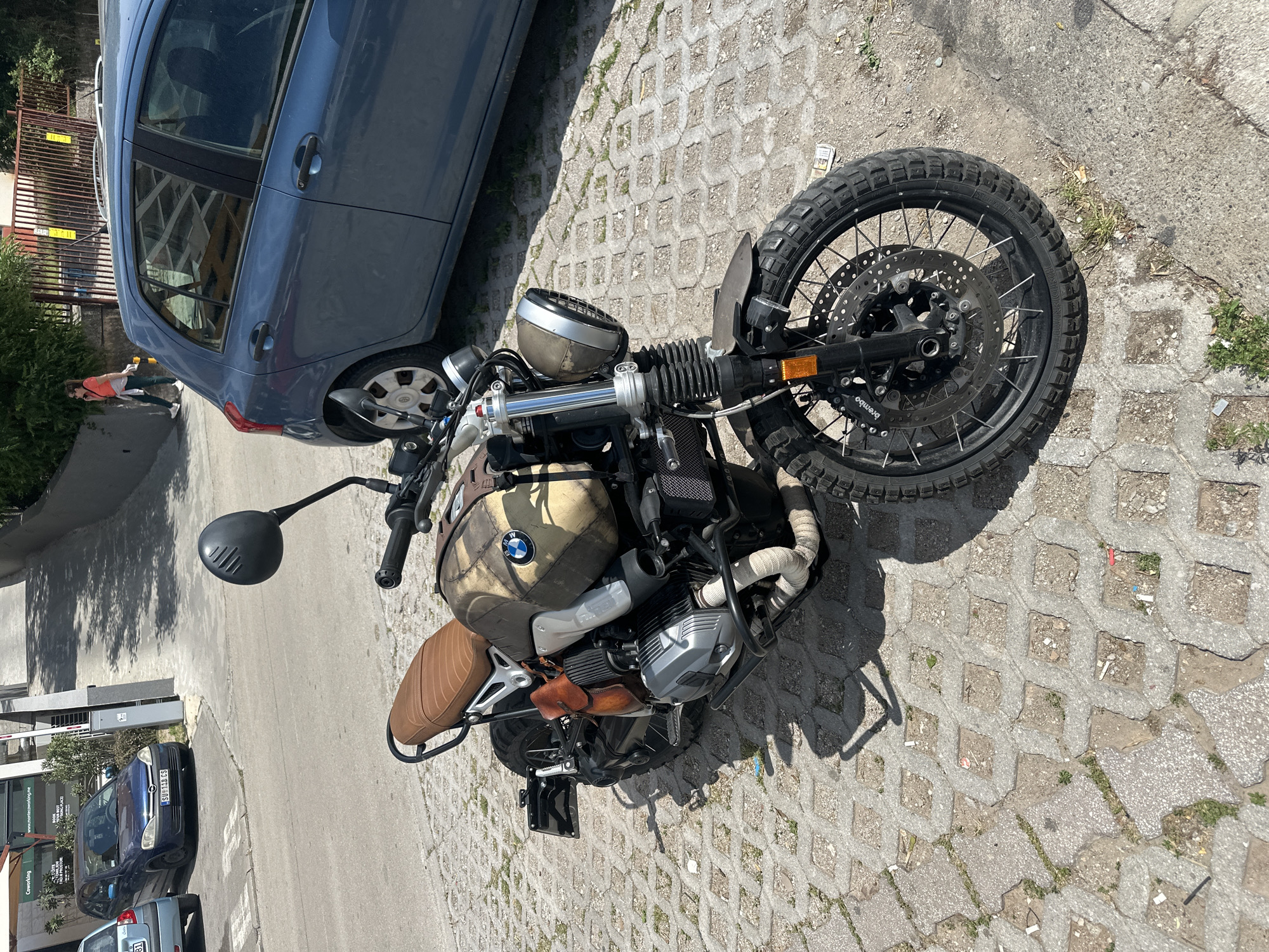 The image depicts a BMW R nineT Scrambler motorcycle parked on a concrete block paved area in the city of Budva, Montenegro. The motorcycle is brown and has a tan seat. It is parked next to a blue car. Another blue car can be seen parked on the street in the background. A person is walking in the background, next to the blue car. The time of day appears to be mid-day, as suggested by the shadows. A building can be seen in the background with green signage.