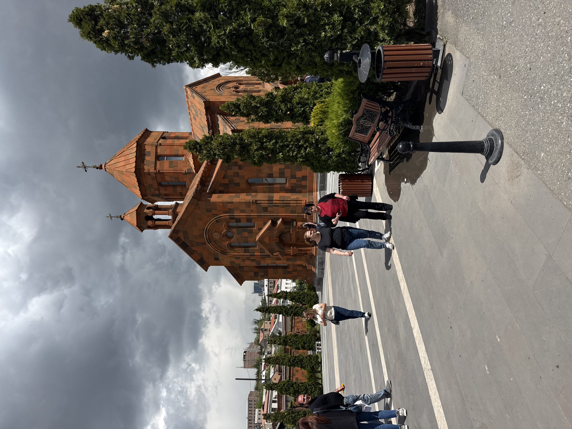 The image depicts Saint Anna Church in Yerevan, Armenia. The church, built of orange stone with a tiled roof, is located on a paved plaza with several people walking on it. There are benches and landscaping features, including trimmed bushes and trees. A dark, cloudy sky is above the church. The city's urban landscape is visible in the background.